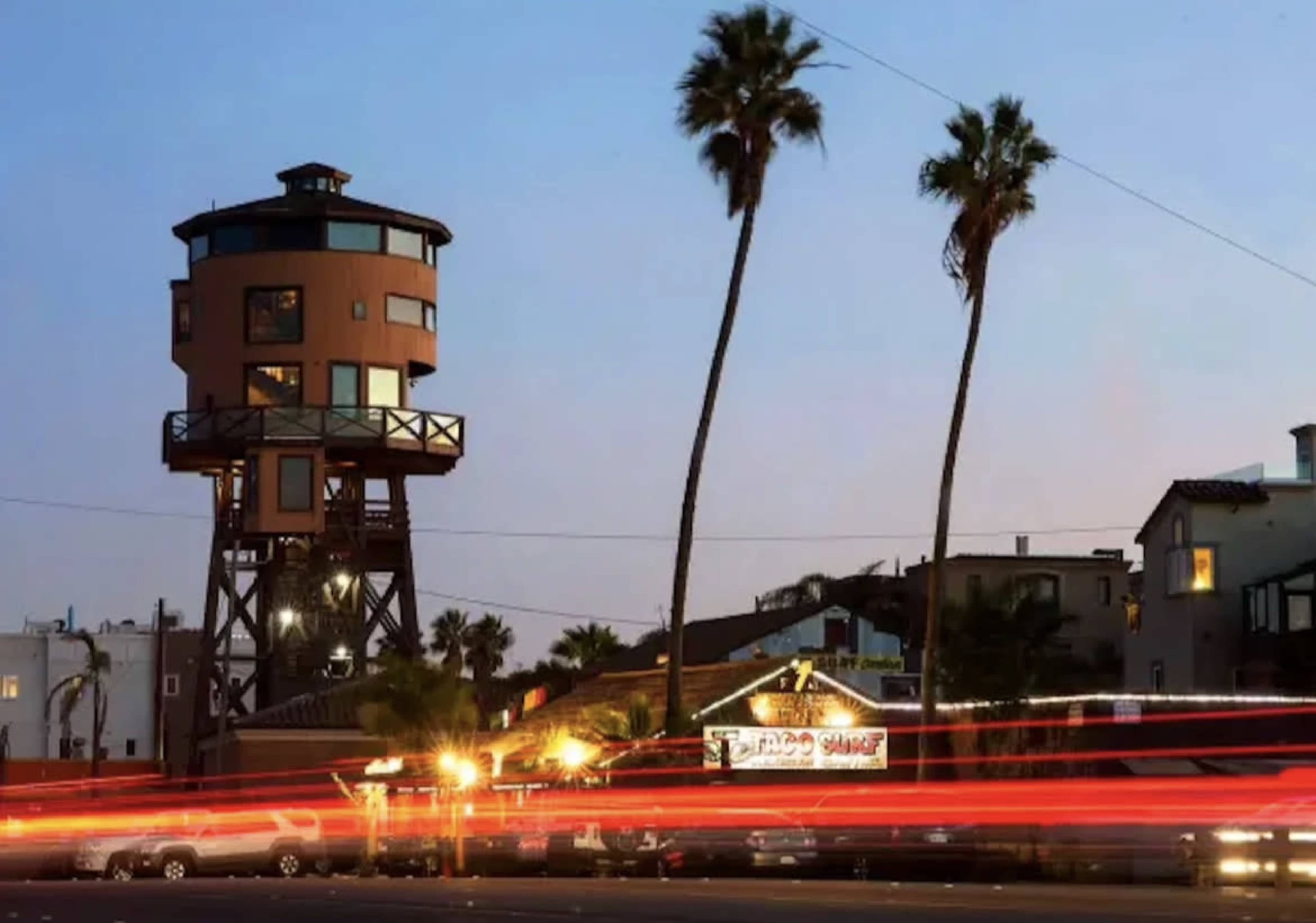 A tall, cylindrical building with a deck stands next to palm trees and a restaurant in a coastal area at dusk.