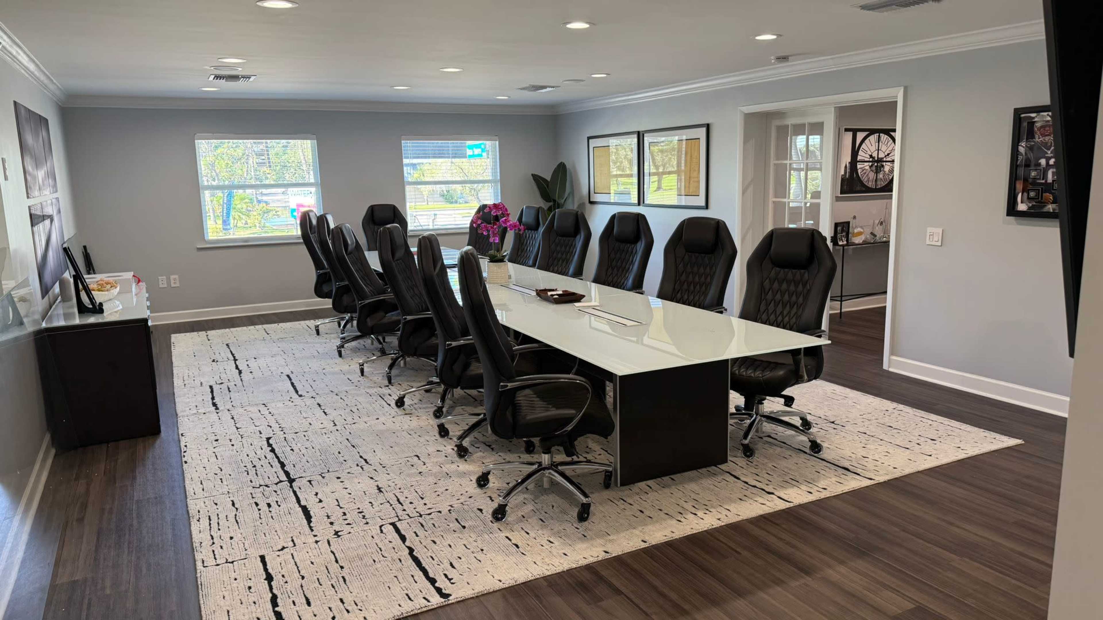 A large, modern conference room features a glossy table surrounded by sleek black chairs, with windows offering natural light and a decorative rug on the floor.