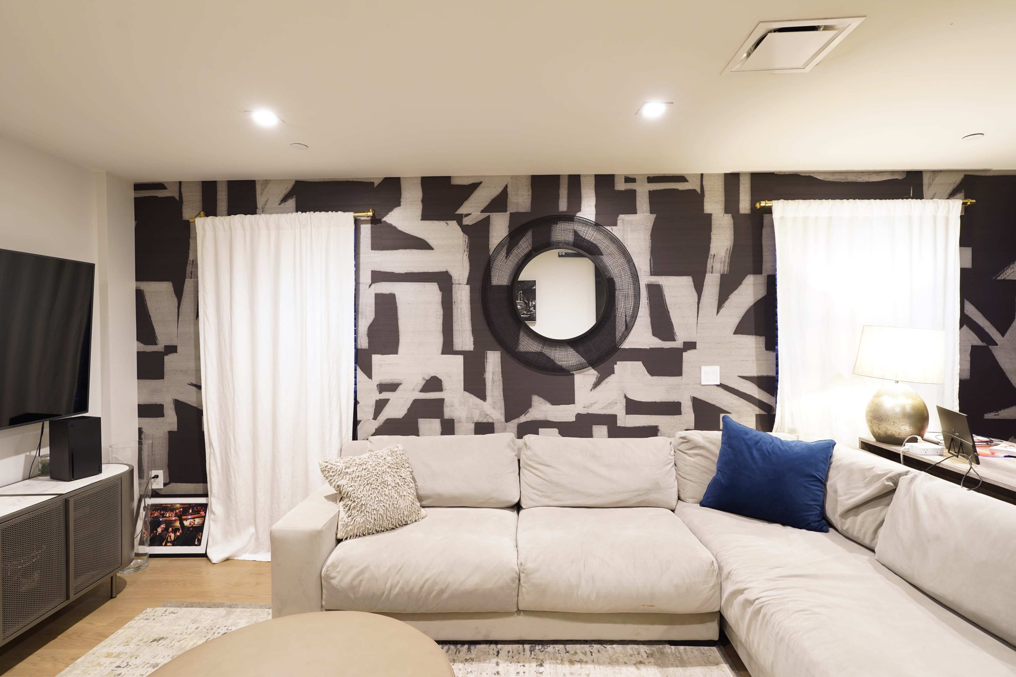 The image shows a modern living room with a light-colored sectional sofa, a round coffee table, and patterned wallpaper, along with large curtains and a television mounted on the wall.