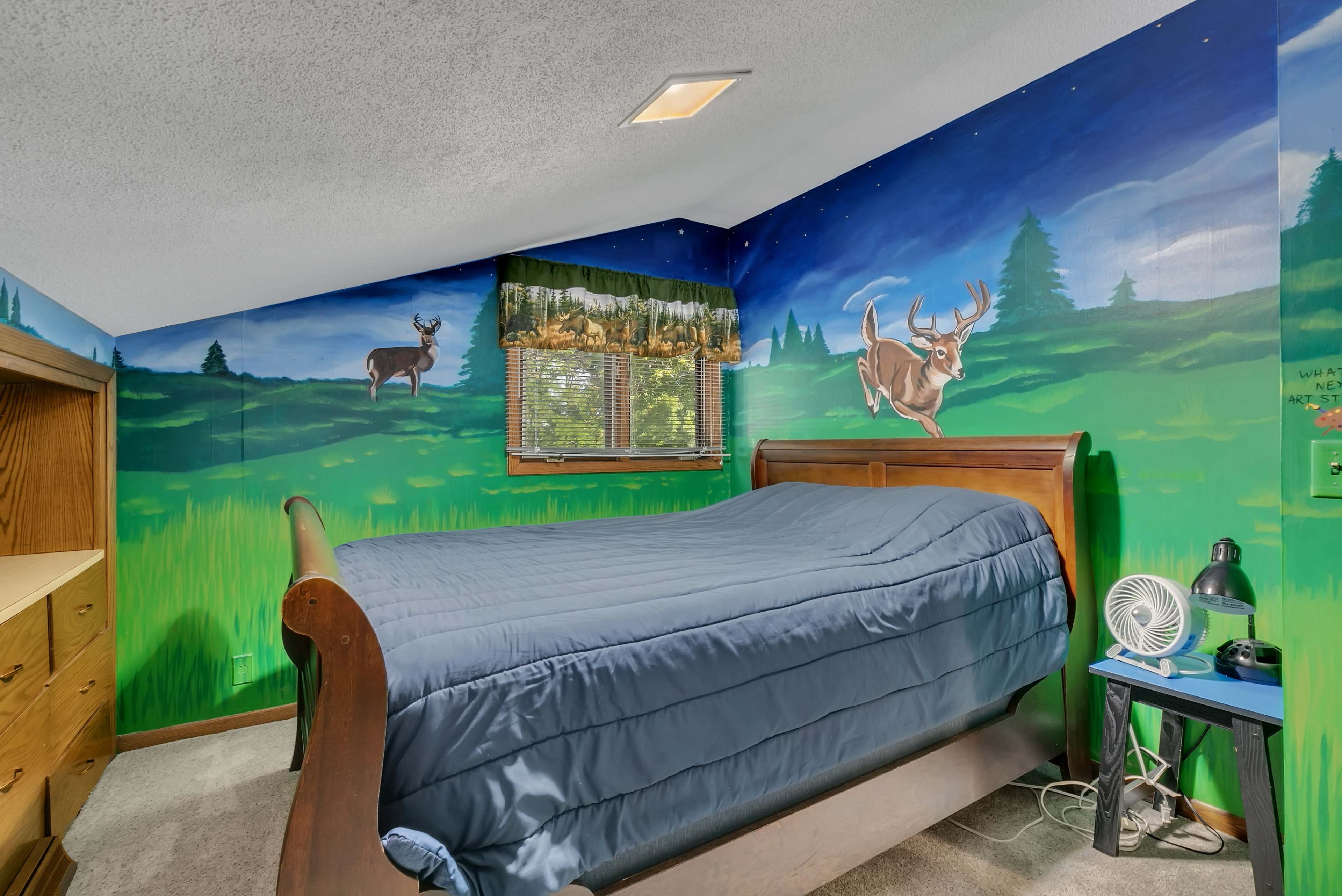 The image shows a bedroom with a wooden bed, a painted landscape mural on the walls featuring deer and trees, and a fan beside the bed.