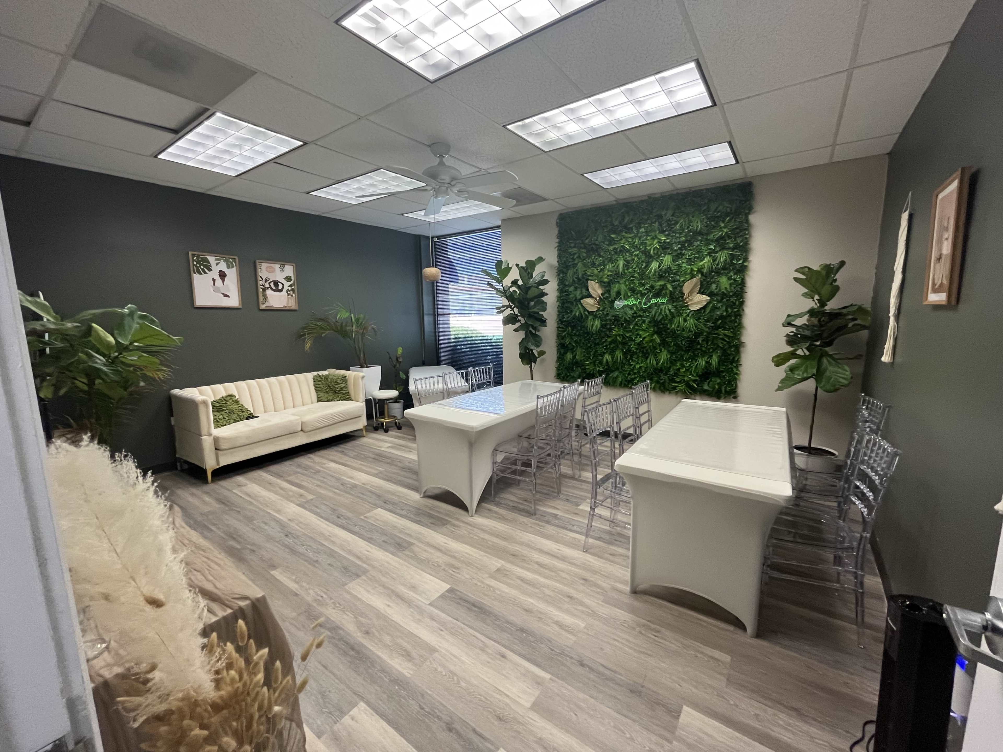 A conference room with a sofa, several transparent chairs, a white table, decorative plants, and a green wall feature.