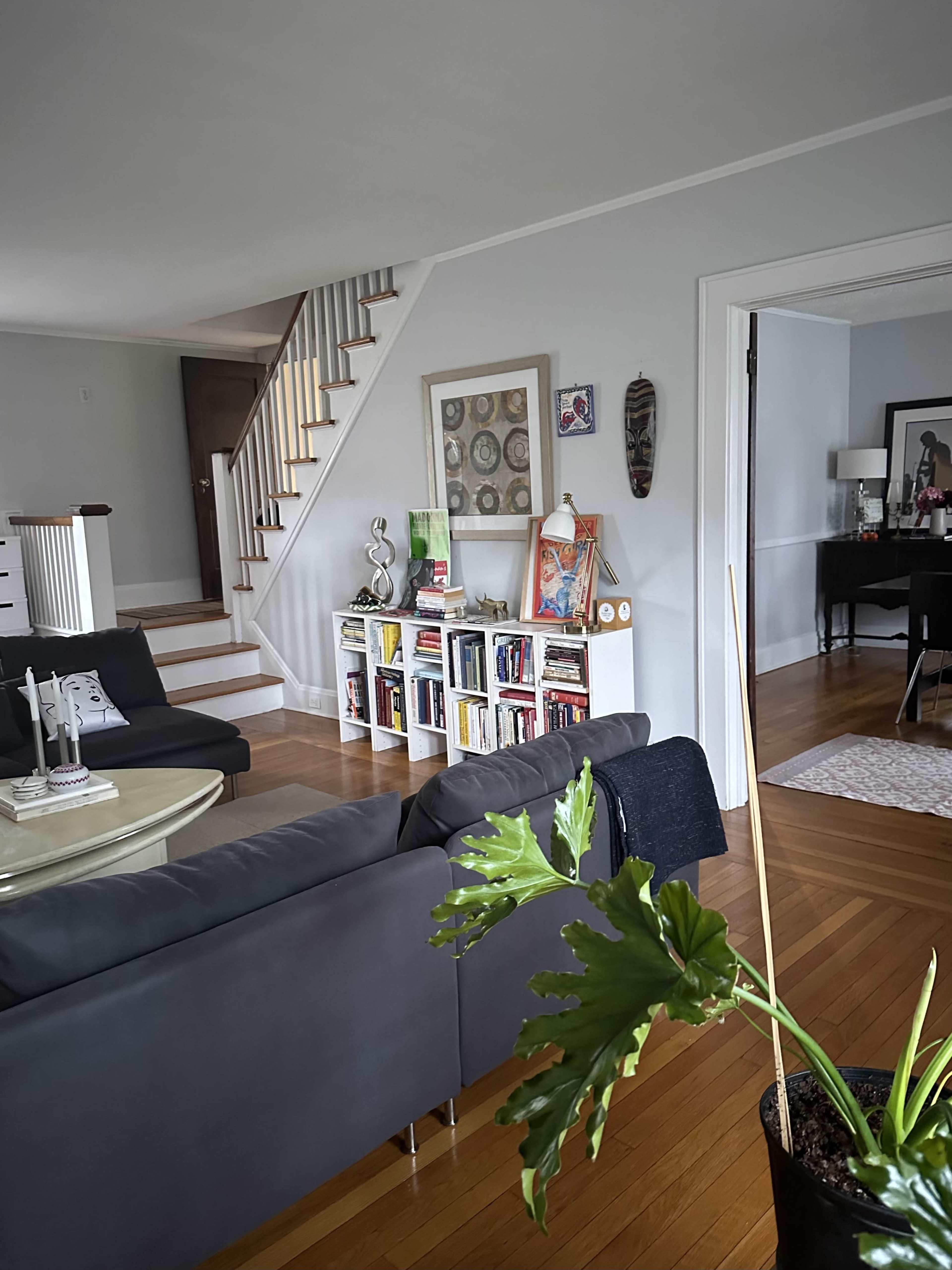 The image shows a living room with a black couch, a plant, a bookshelf filled with books, and a staircase in the background.