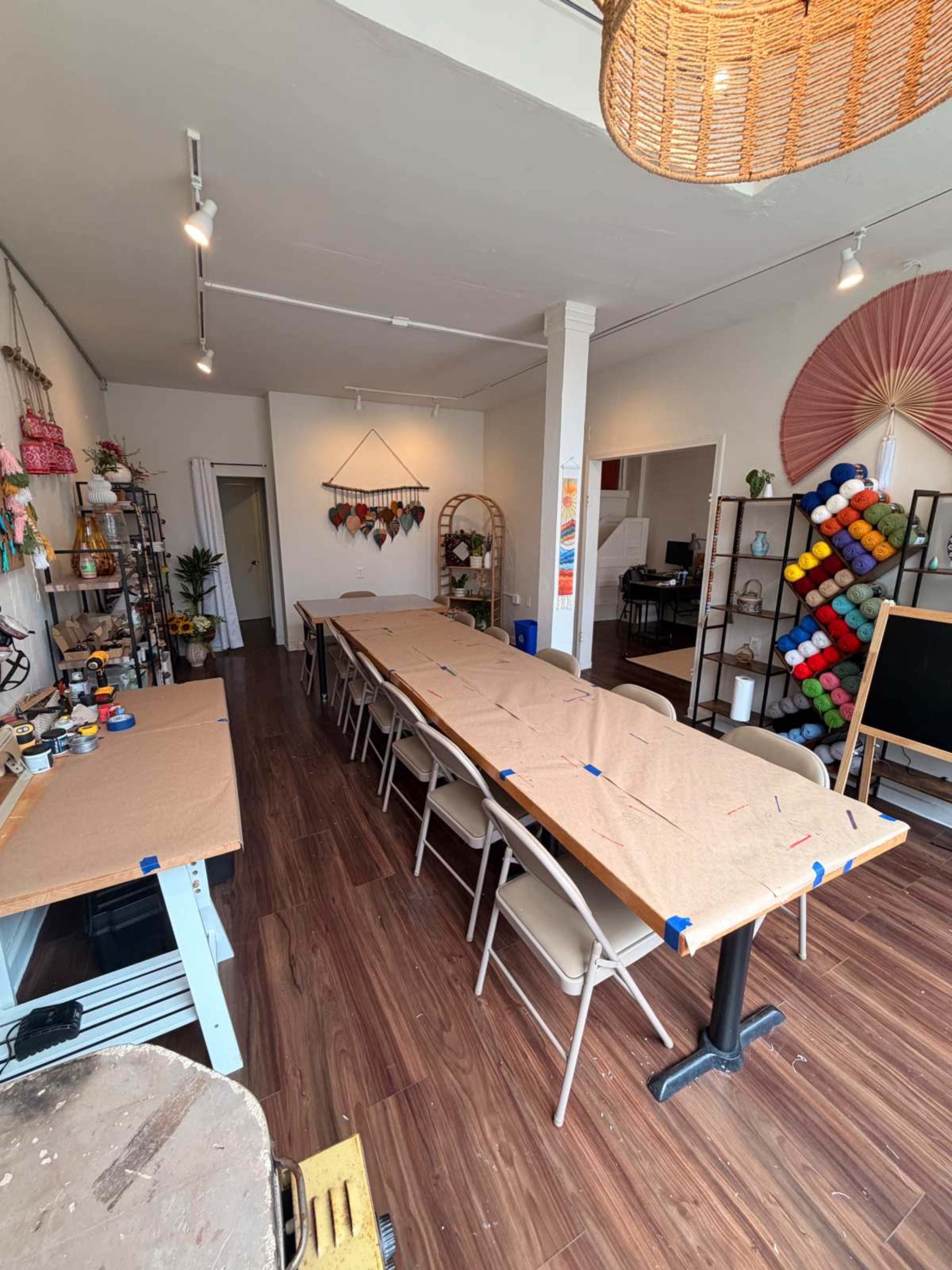 The image shows a spacious room with several tables covered in brown paper, arranged for an event, and decorated with plants and colorful craft supplies along the walls.