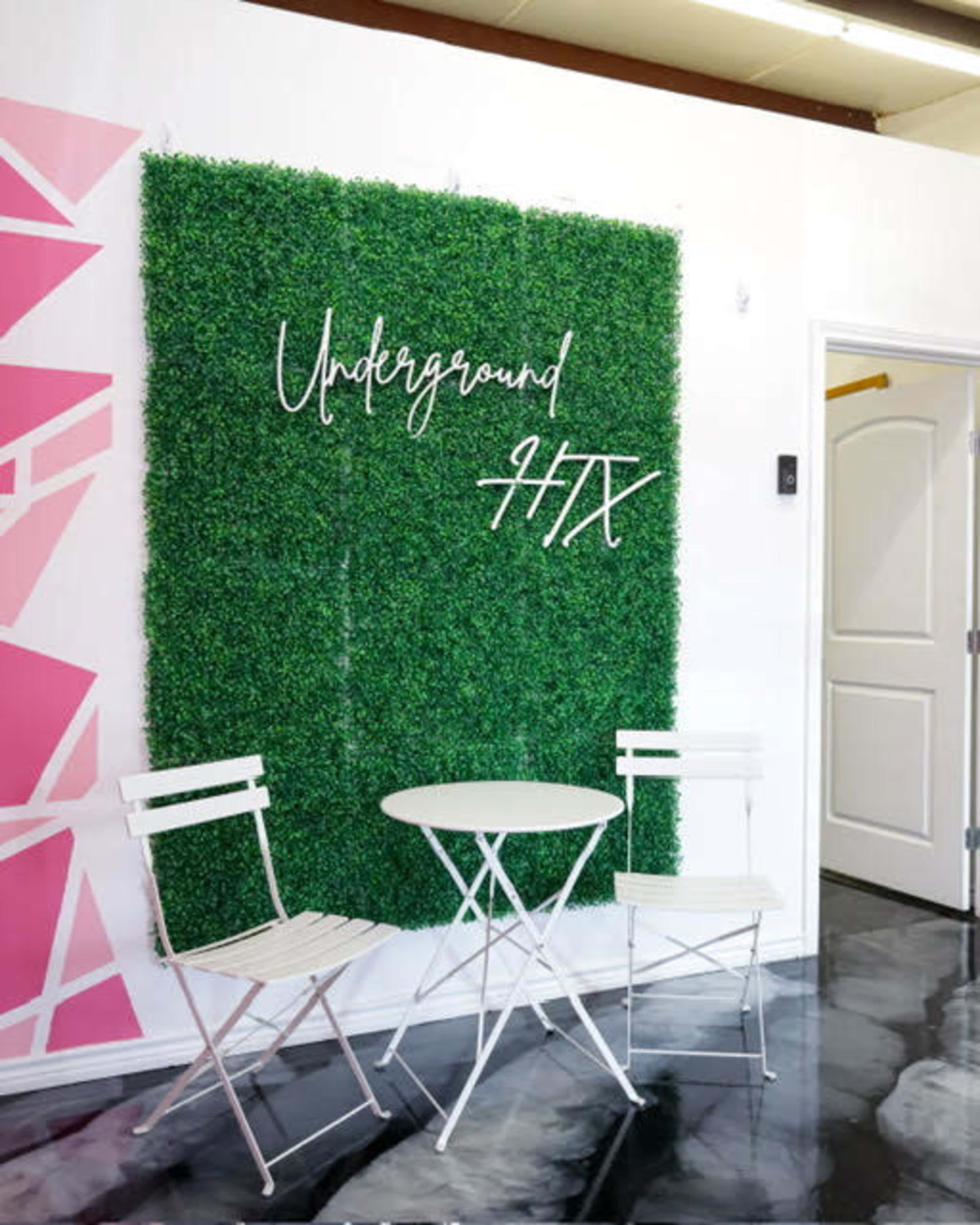 The scene features a small sitting area with a round white table and two white chairs in front of a green artificial grass wall, with the text "Underground HTX" displayed on it.