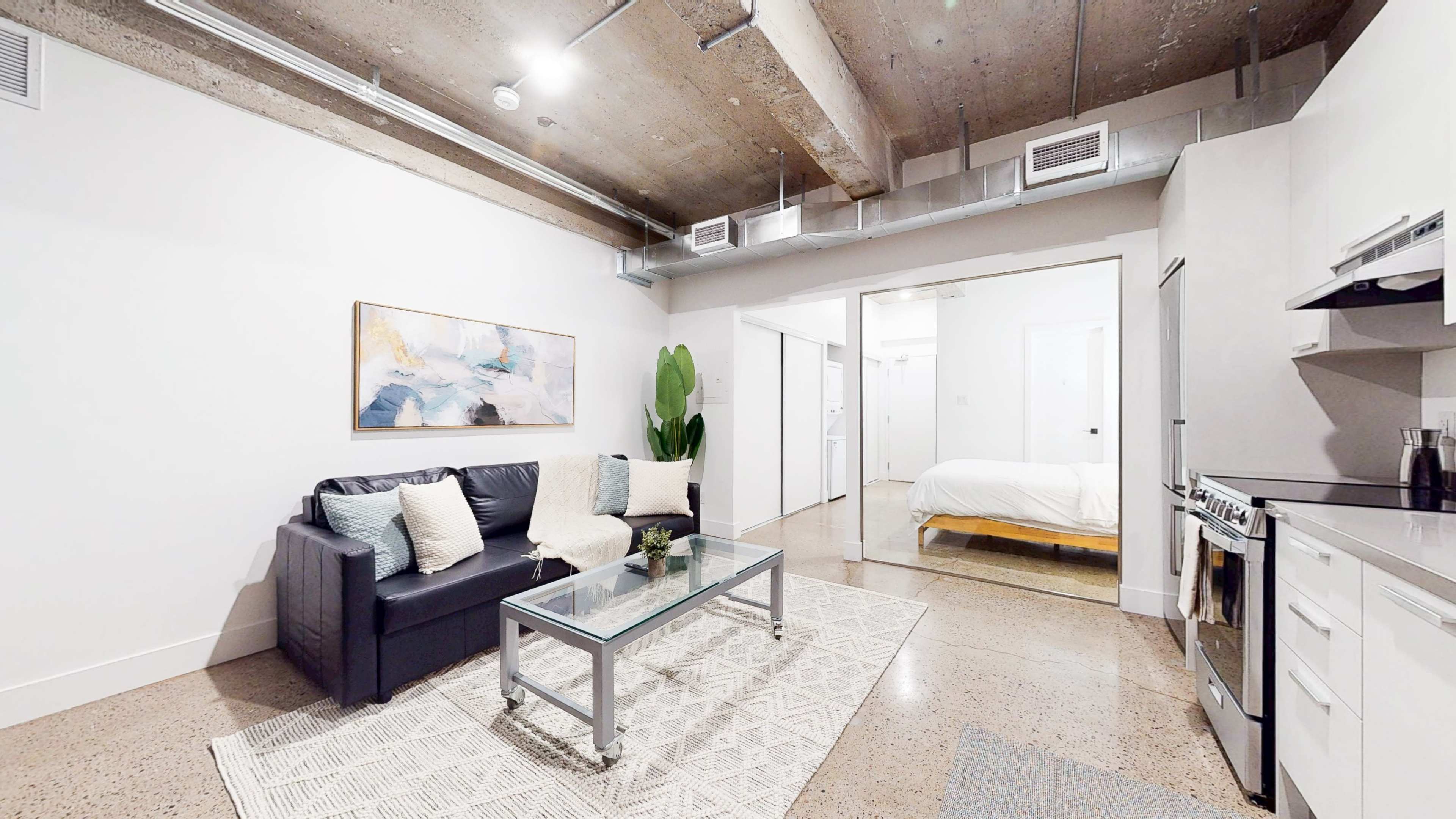 The image shows a modern living space with a black sofa, a glass coffee table, and an open kitchen area, featuring concrete ceilings and flooring.