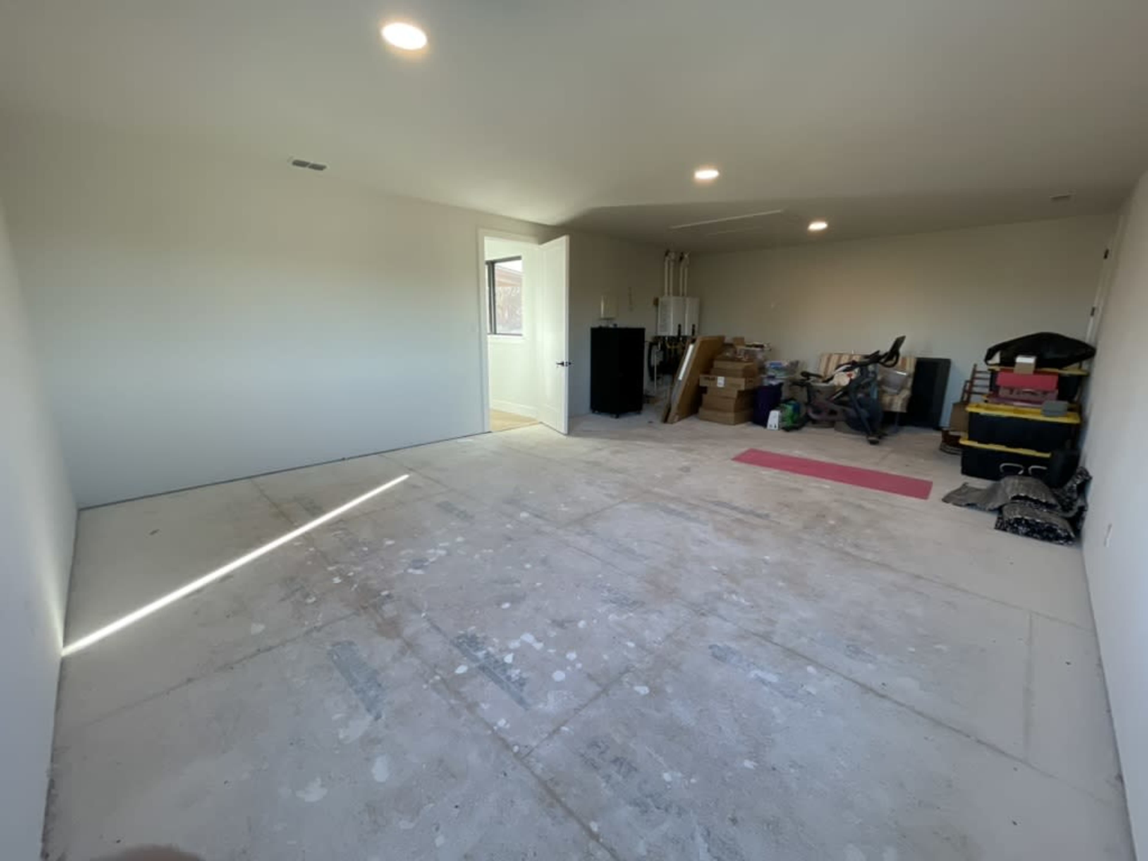 The interior of a bare, unfinished room features a concrete floor, white walls, and a doorway leading to another space, with various boxes and equipment placed along the sides.