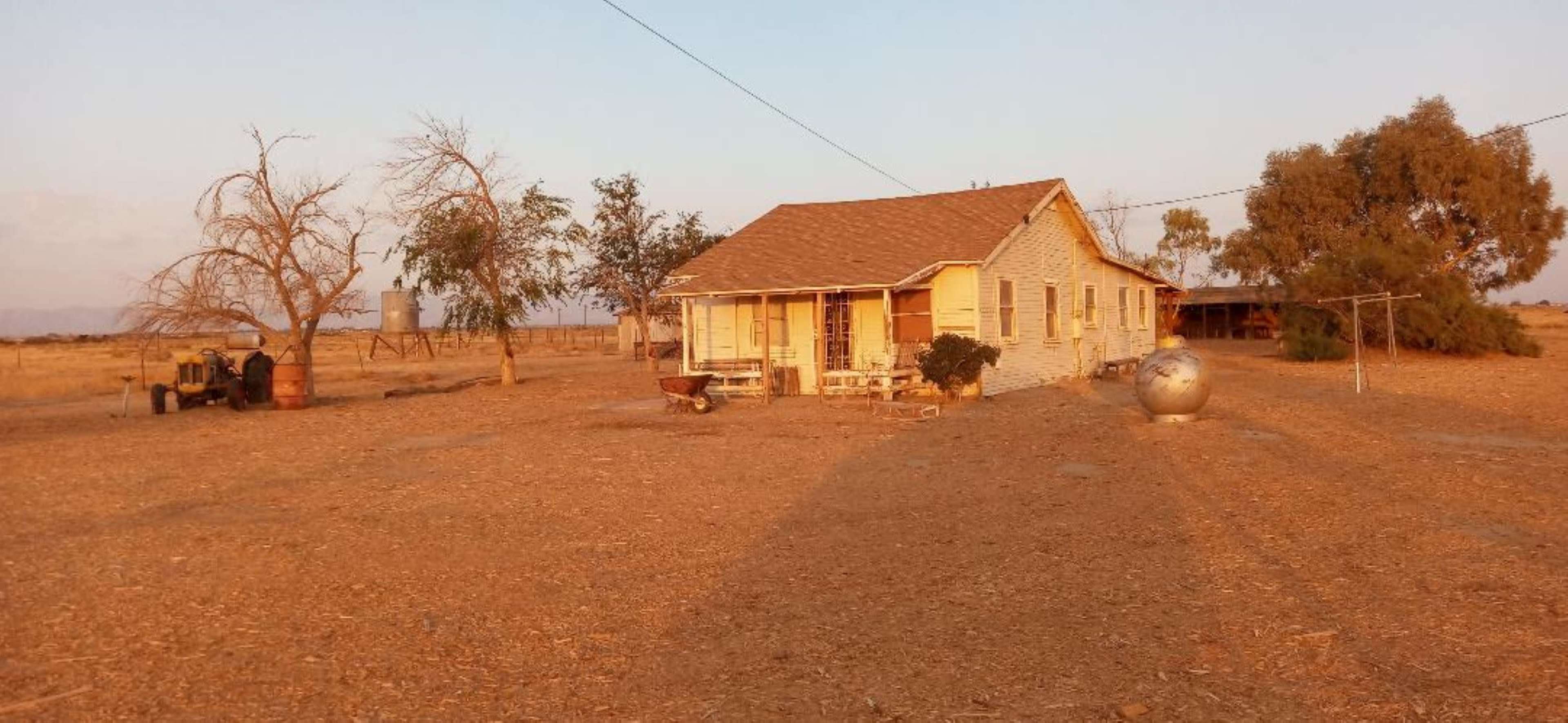 A single-story house with a front porch is situated in a barren landscape surrounded by sparse trees and agricultural equipment.