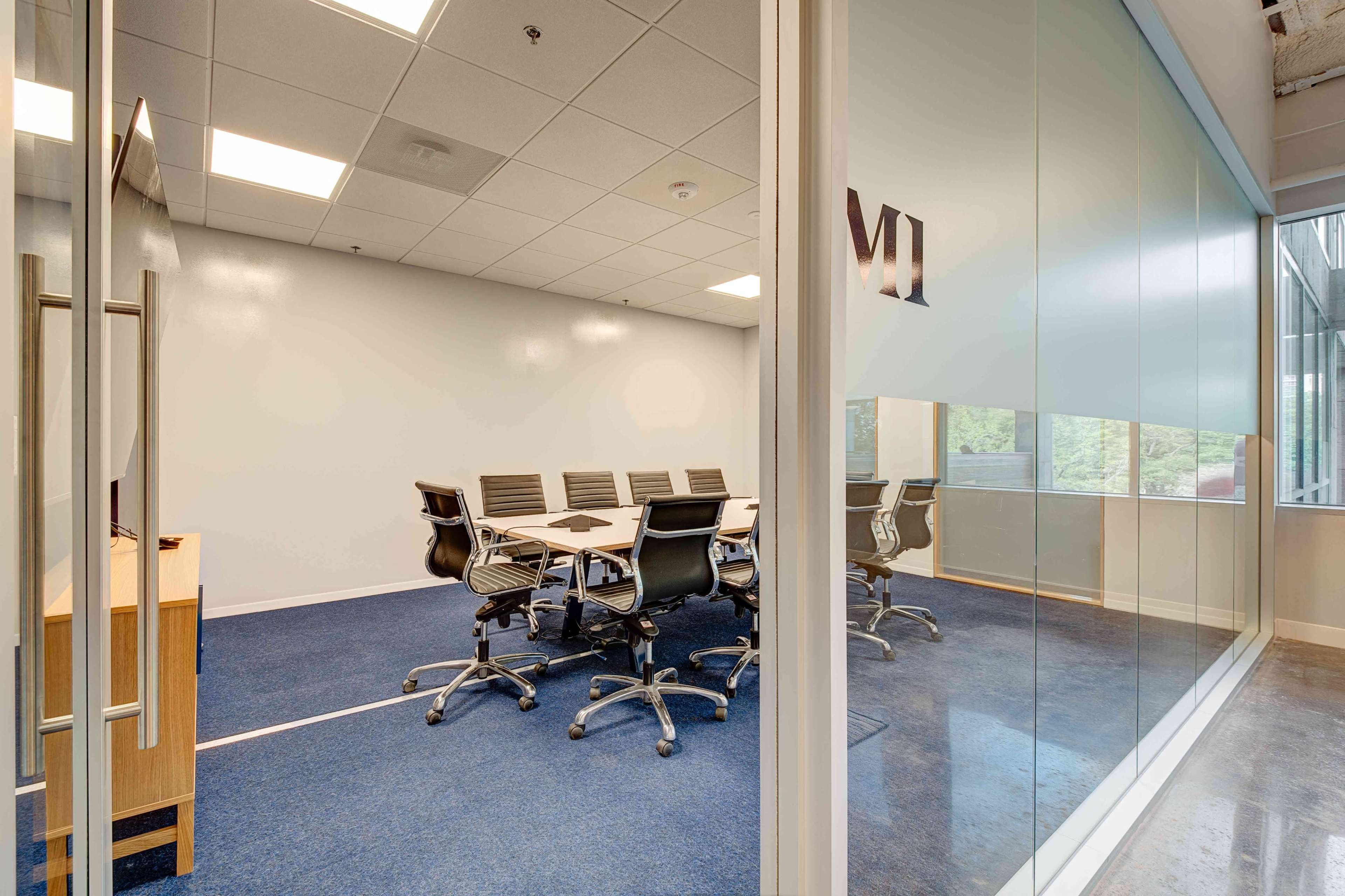 A modern conference room with a glass wall features a large table surrounded by black chairs and a television on one side.