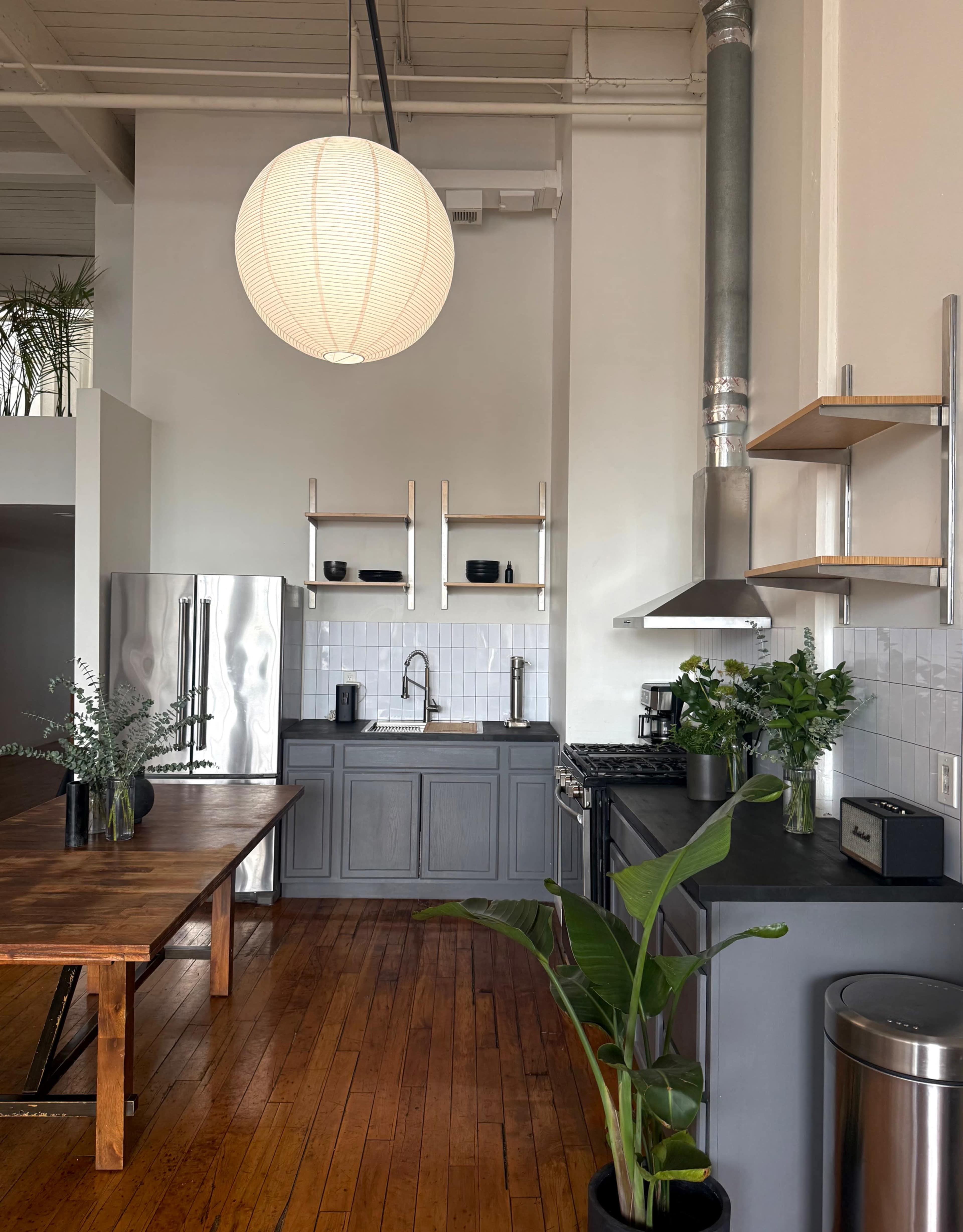 The image shows a modern kitchen with a grey countertop, stainless steel appliances, wooden floors, and a large pendant light hanging from the ceiling.