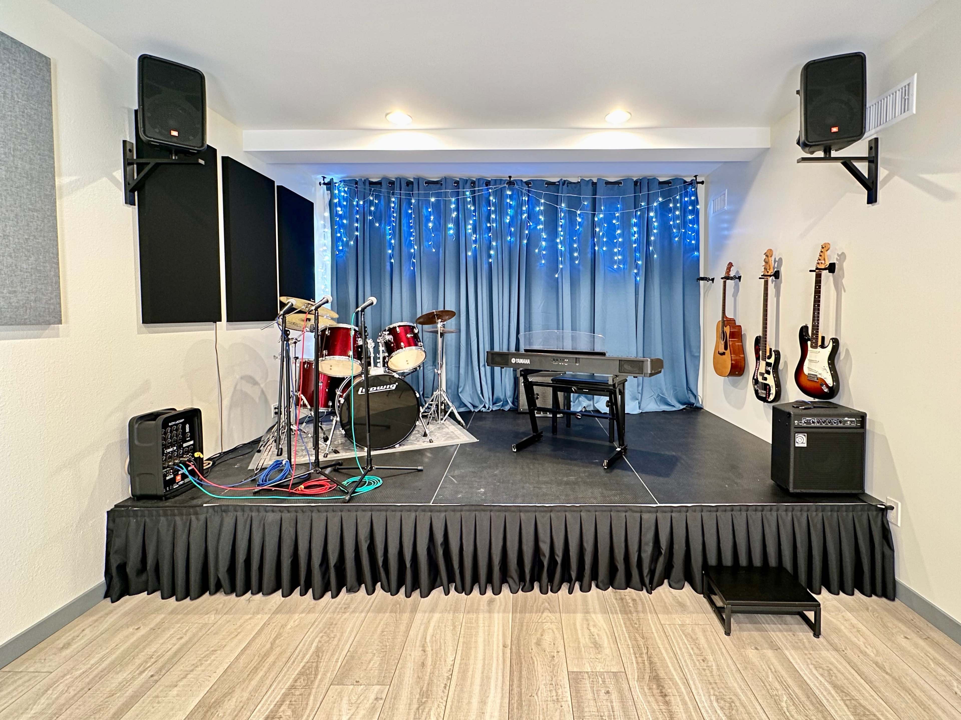 A small stage setup featuring a drum kit, a keyboard, and several electric guitars mounted on the wall, with blue curtains and string lights in the background.