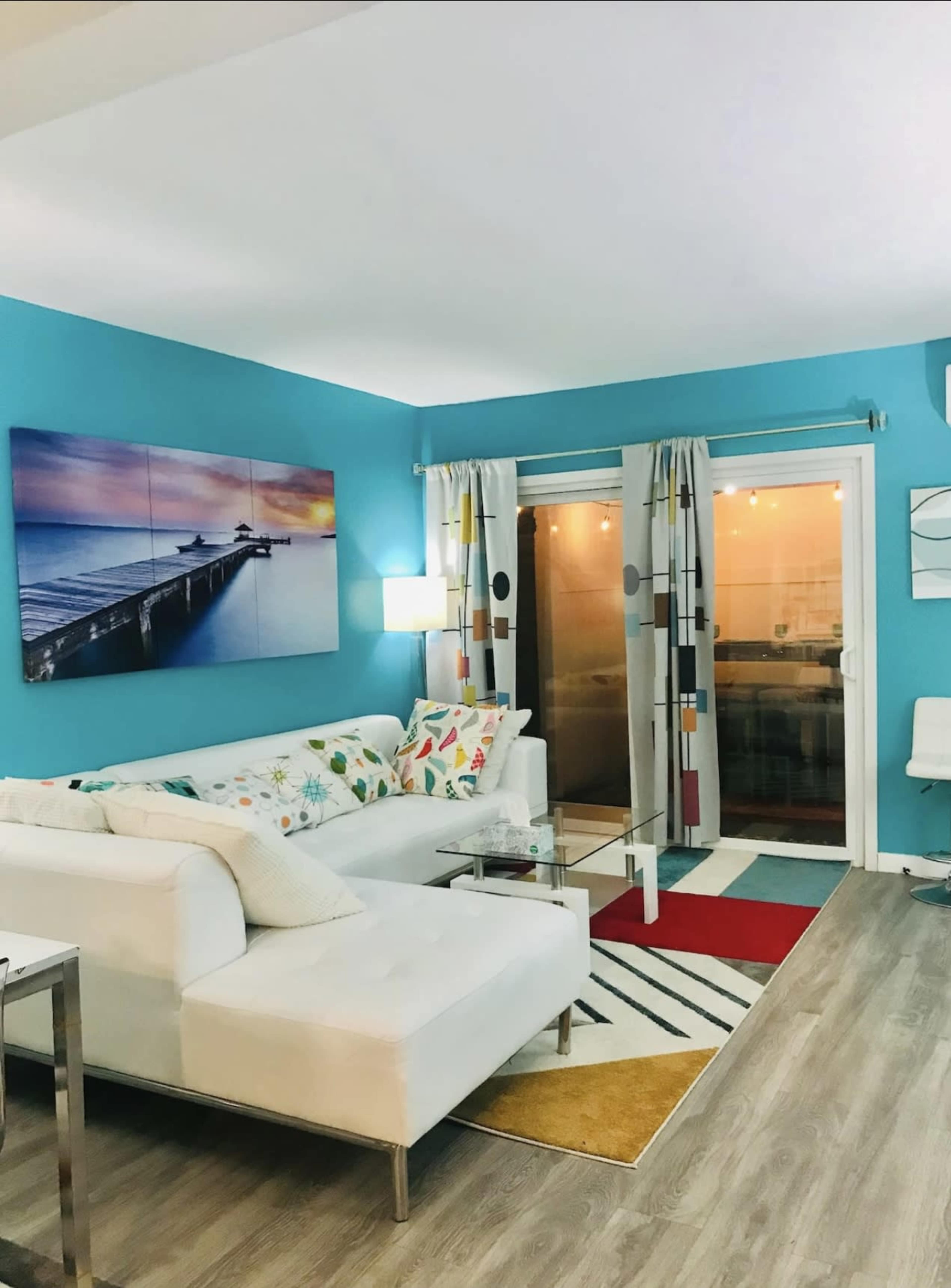 The image shows a brightly colored living room with a white sectional sofa, a glass coffee table, and a large piece of wall art featuring a pier at sunset.