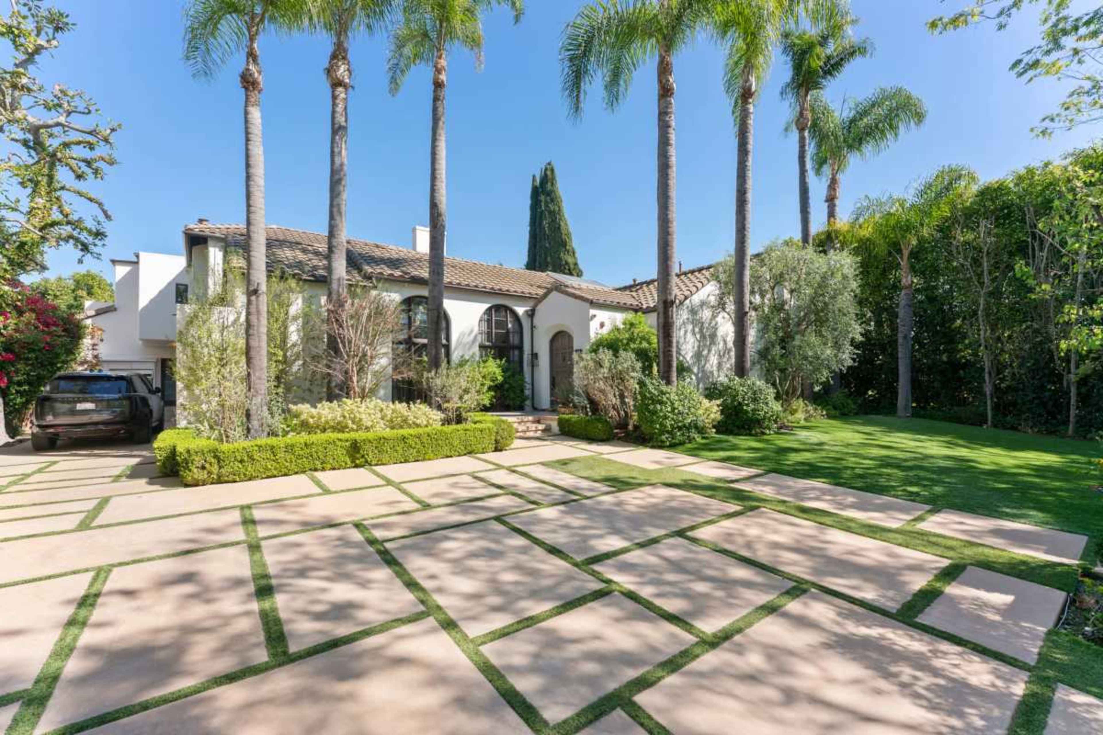 The image shows a modern single-story house surrounded by palm trees and well-maintained landscaping, with a spacious driveway featuring a grid pattern of concrete and grass.