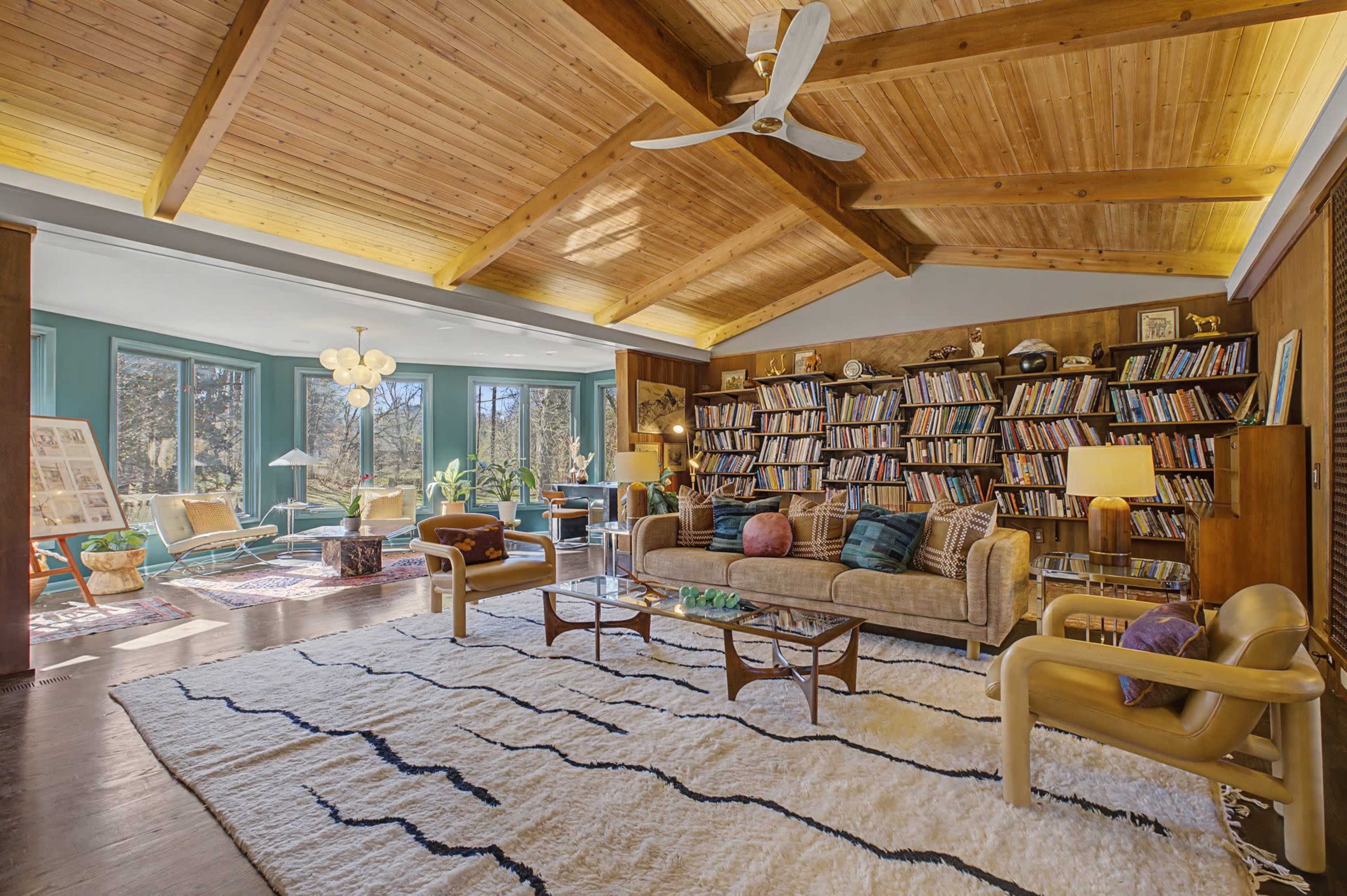 The image shows a spacious living room with a wooden ceiling, a large bookshelf filled with books, various seating arrangements, and large windows letting in natural light.