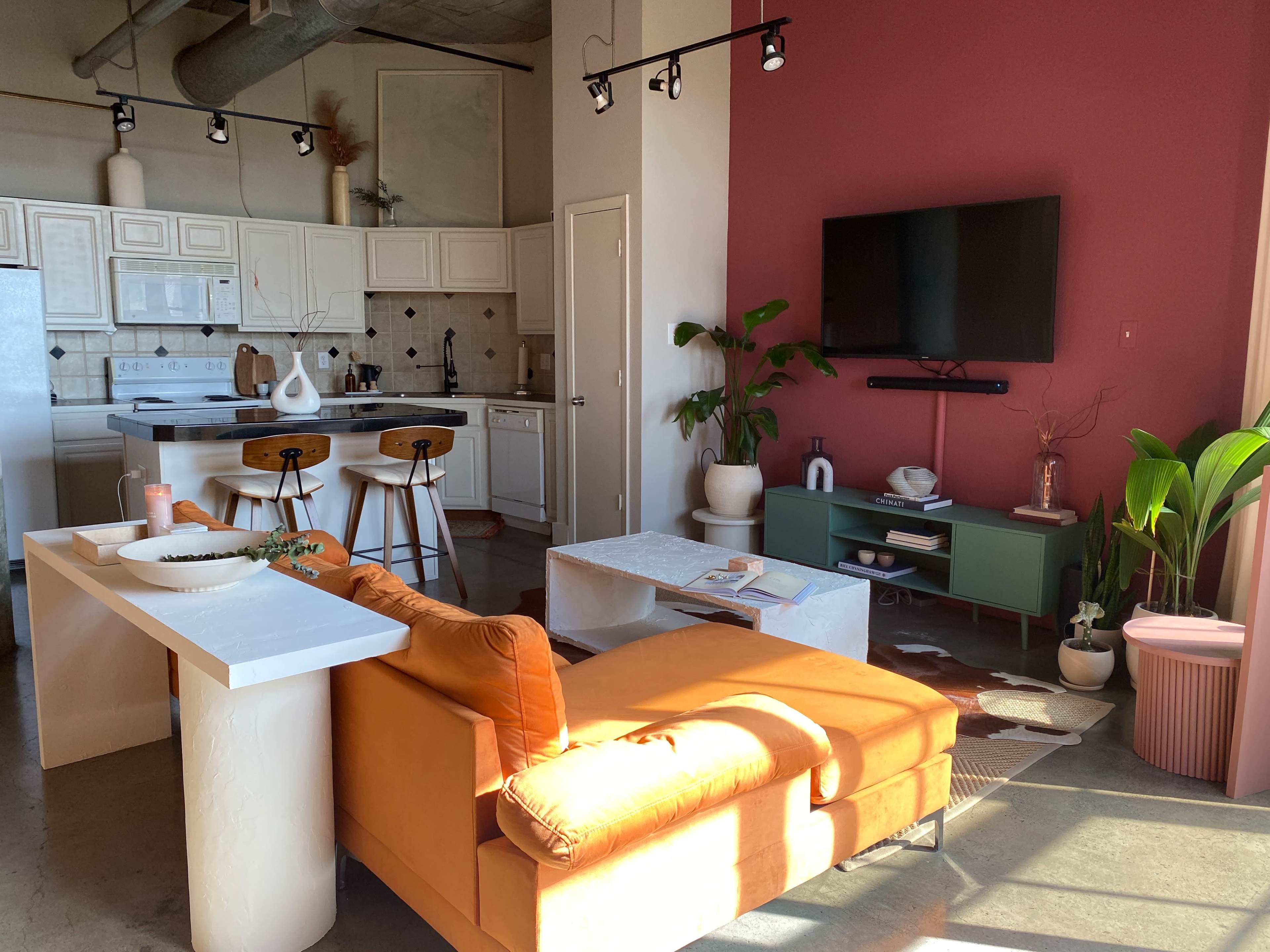 A cozy living space features an orange sofa, a modern kitchen area, and a wall-mounted television surrounded by plants and decorative furniture.