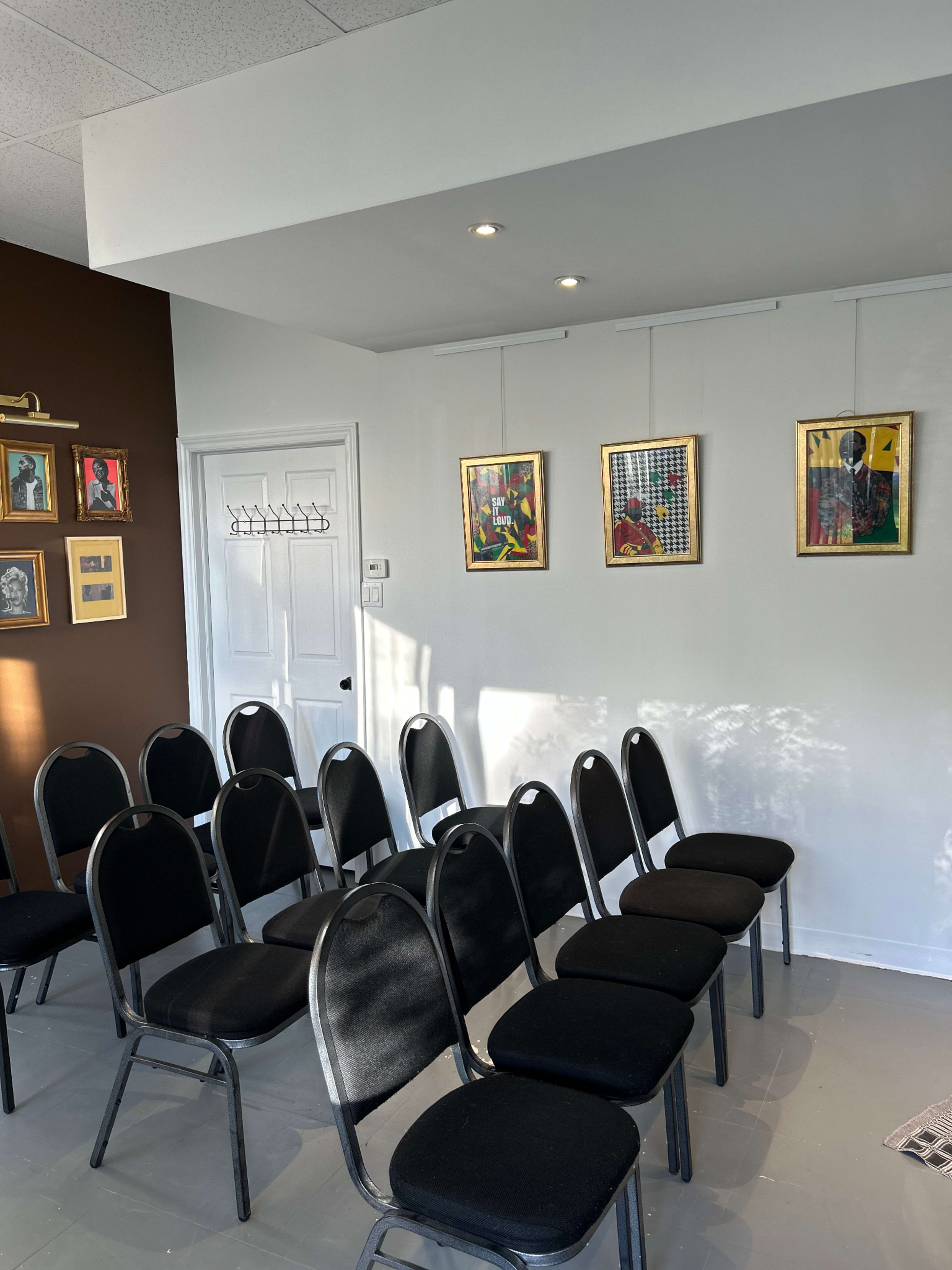 The image shows a room with several rows of black chairs arranged in front of a wall displaying framed artwork.