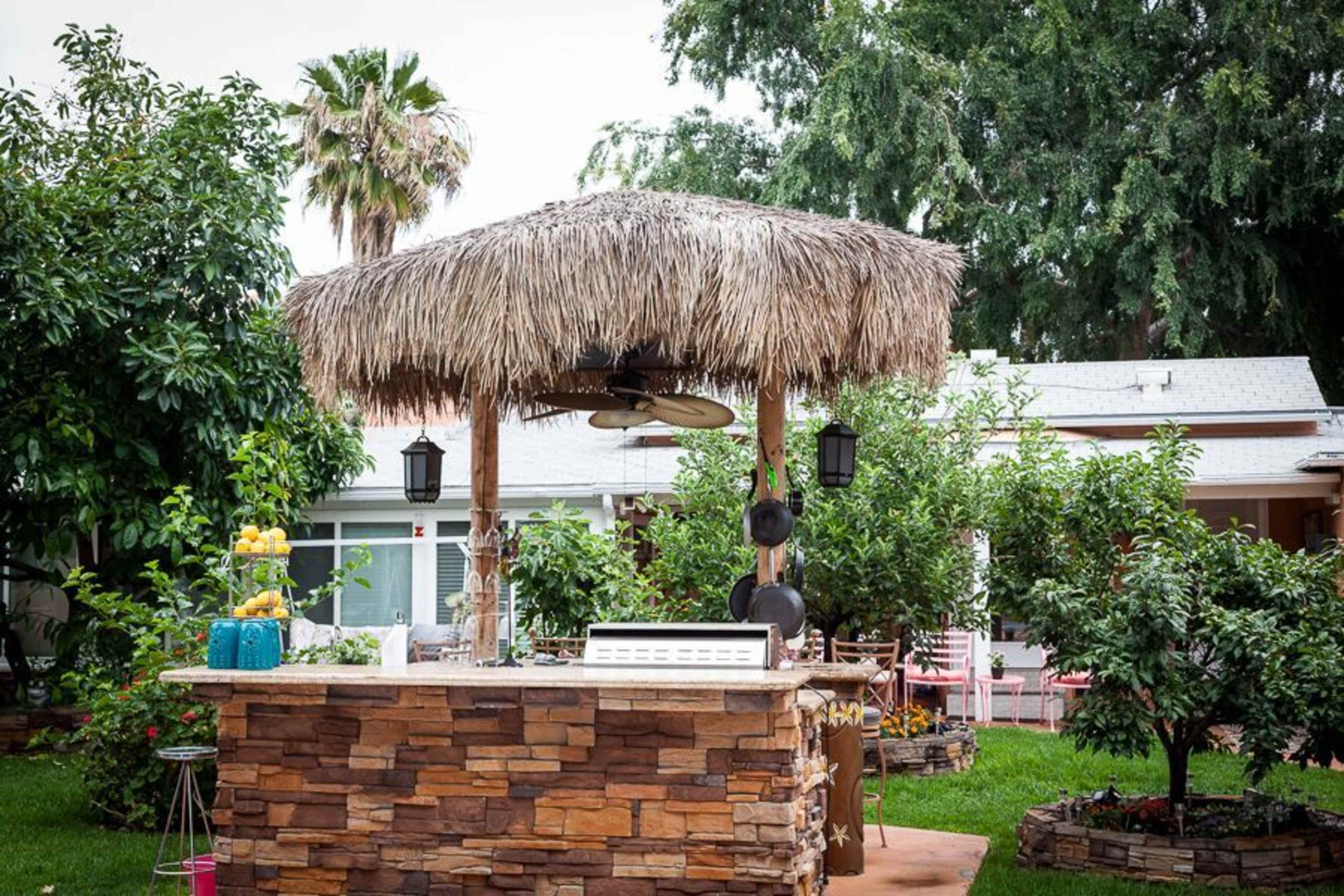 A thatched-roof outdoor bar with stone walls is surrounded by greenery in a backyard.