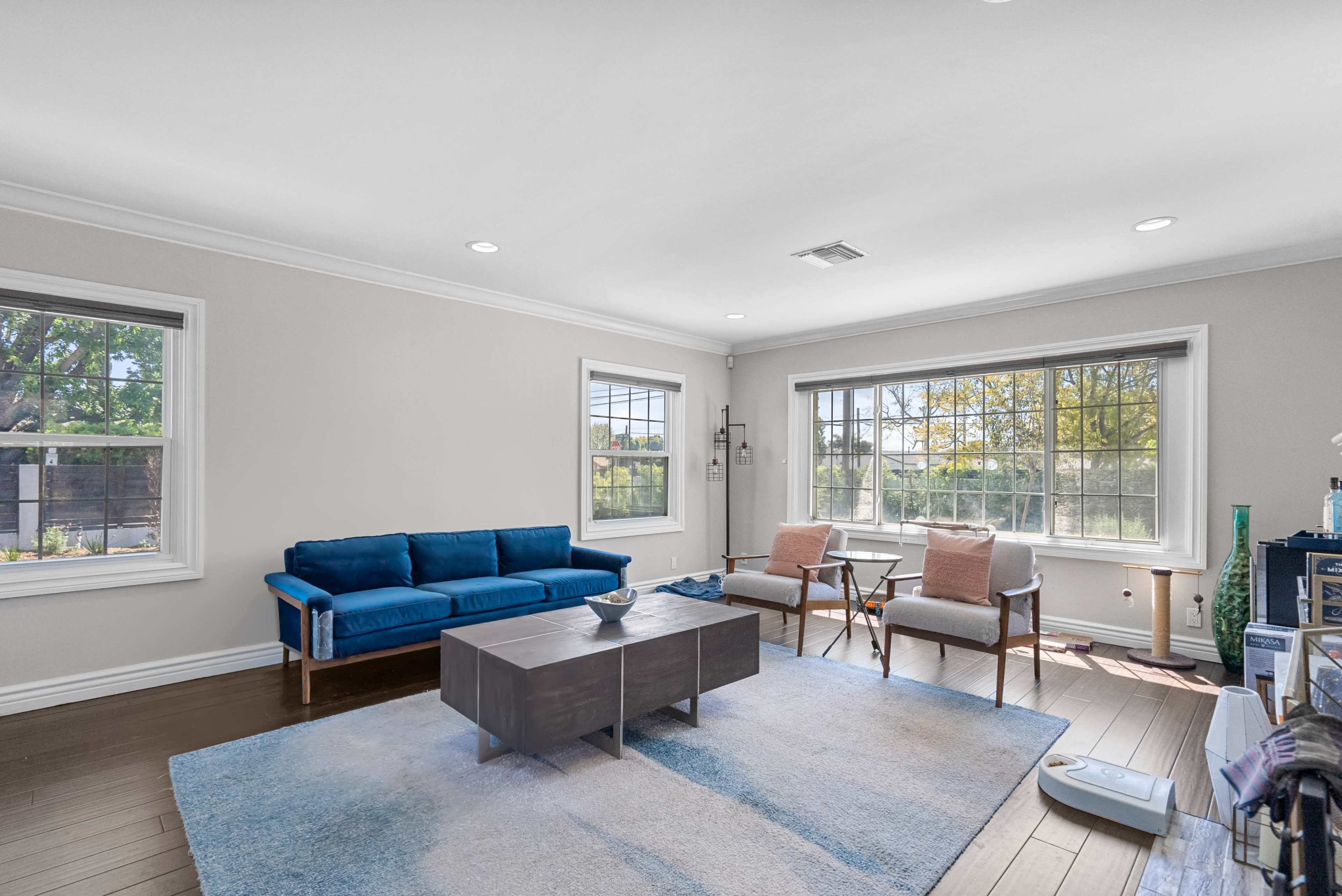 The image shows a bright living room with a blue sofa, two armchairs, a coffee table, and large windows allowing natural light.
