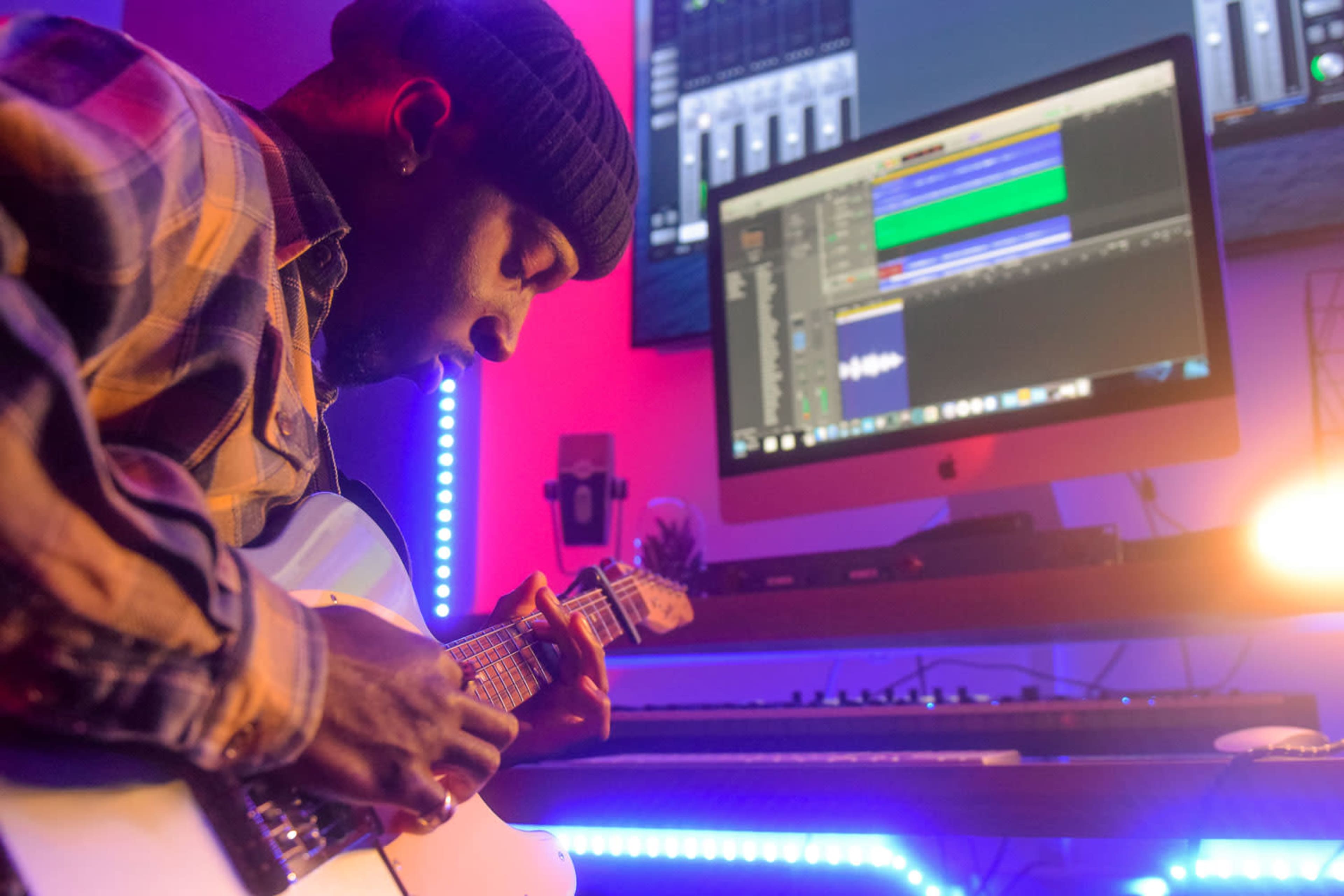 A musician plays an electric guitar in a studio setting with a computer displaying audio software in the background.