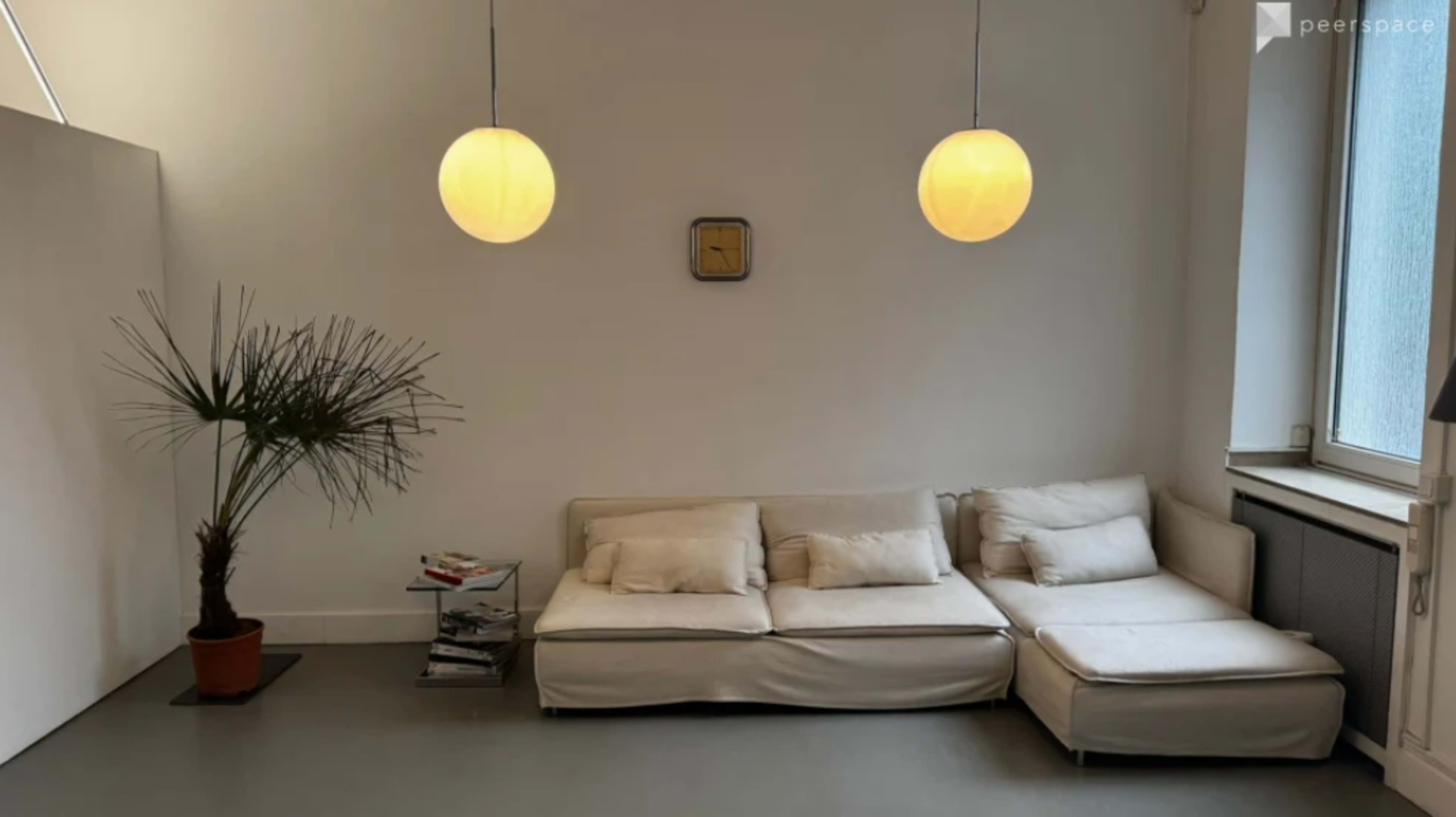 The image shows a minimalistic living space with a light-colored couch, a small table with magazines, a palm plant in a pot, and two pendant lights hanging from the ceiling.