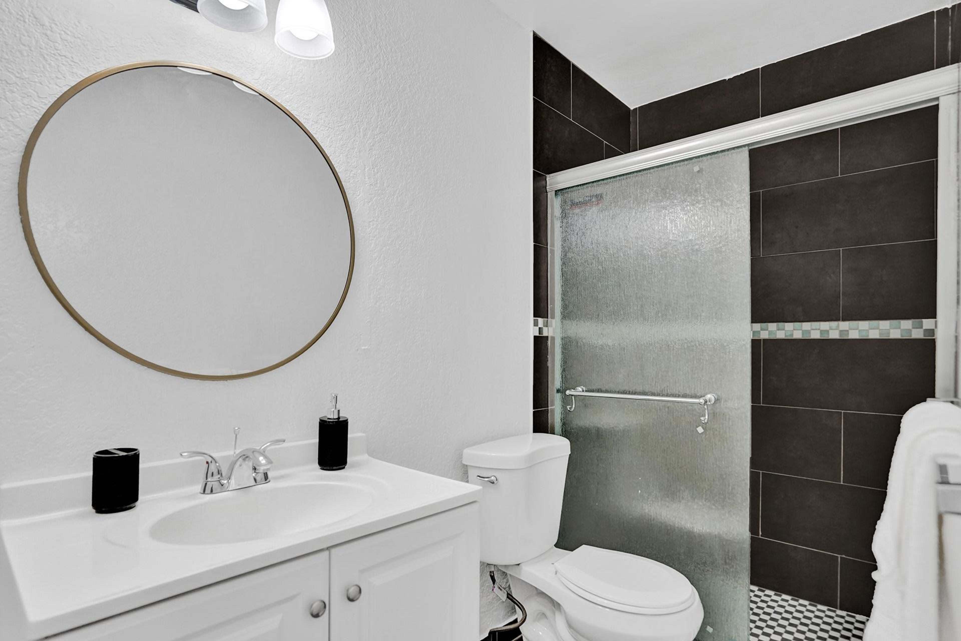 A modern bathroom featuring a circular mirror above a white sink, a toilet, and a glass shower enclosure with dark tiles.