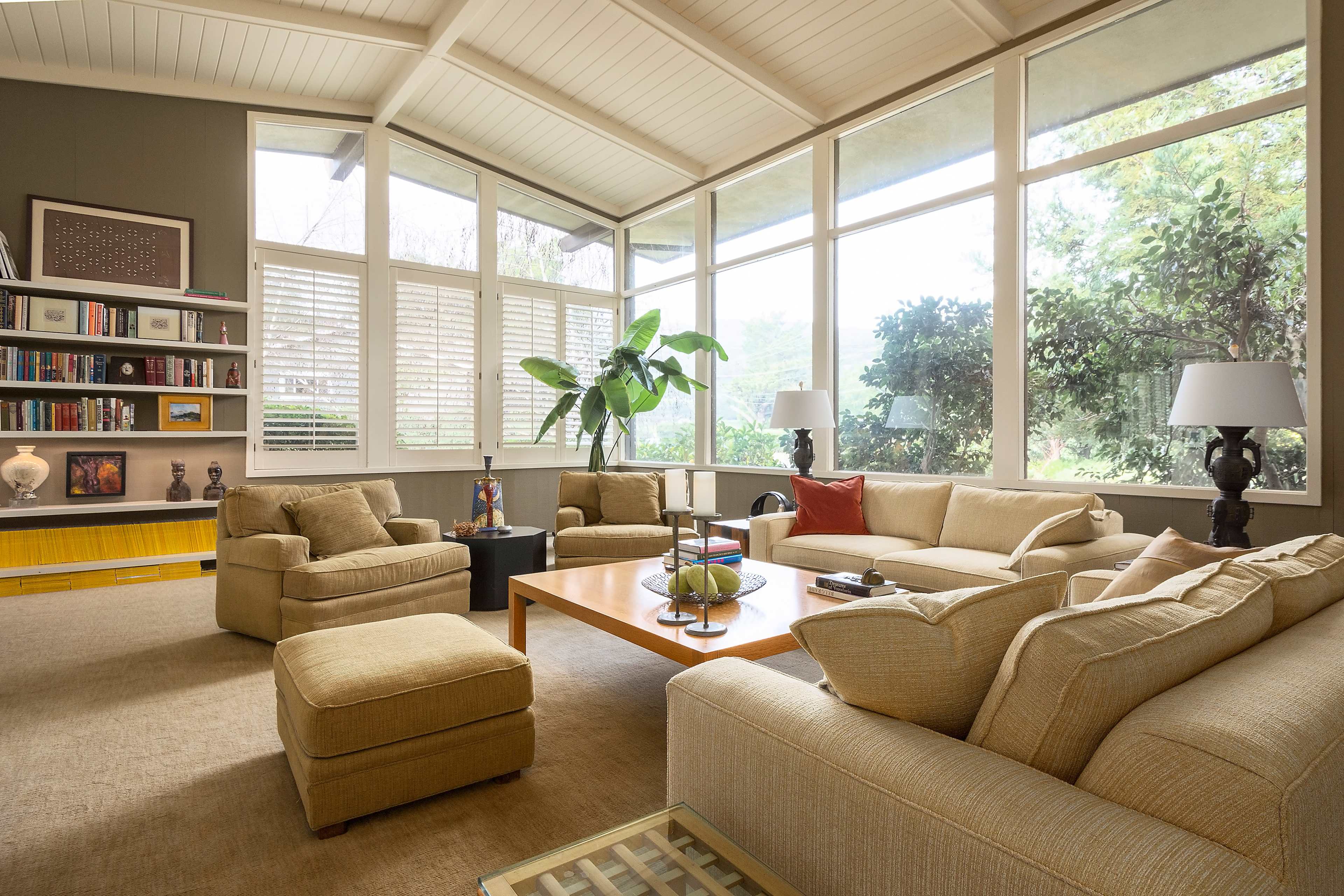 A bright, modern living room with large windows, comfortable beige sofas, and a coffee table surrounded by armchairs.