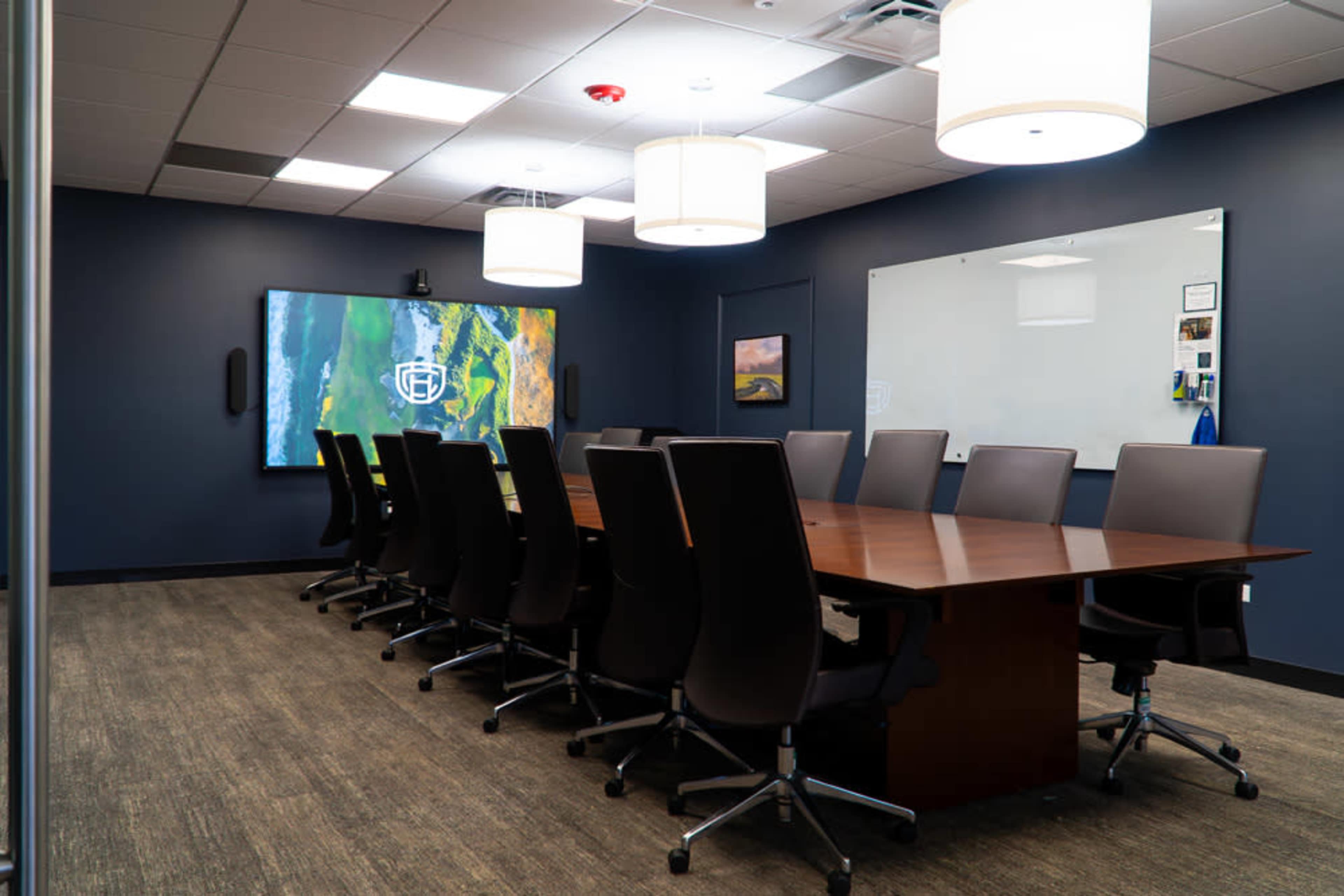 The image shows a modern conference room featuring a large wooden table surrounded by black chairs, with a screen displaying a colorful landscape on one wall and a whiteboard on another.
