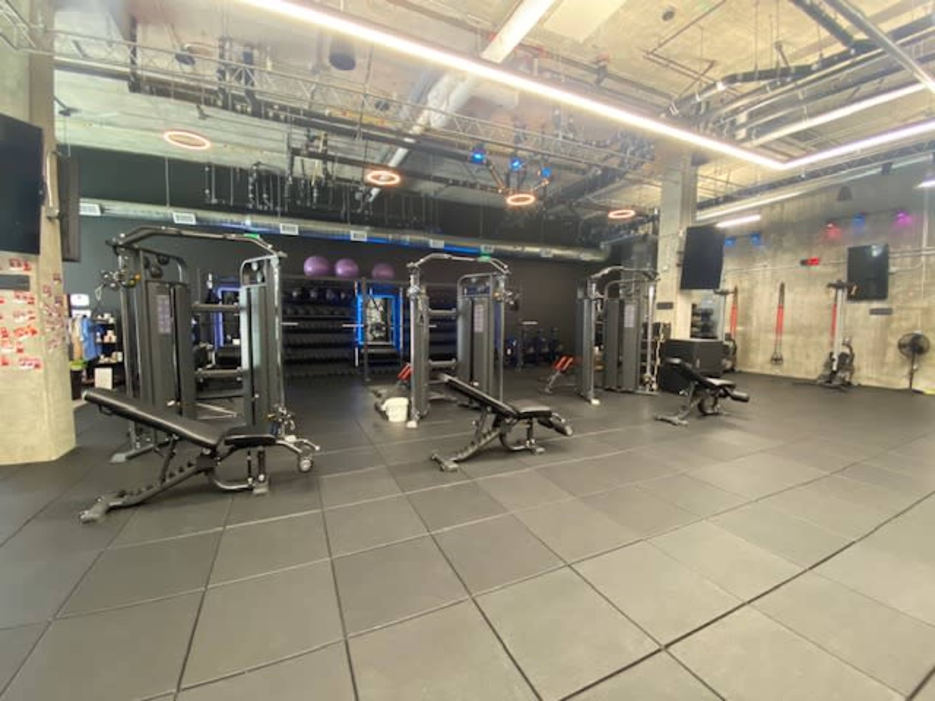 The image shows a modern gym space with several weight machines, benches, and exercise equipment on a rubberized floor.