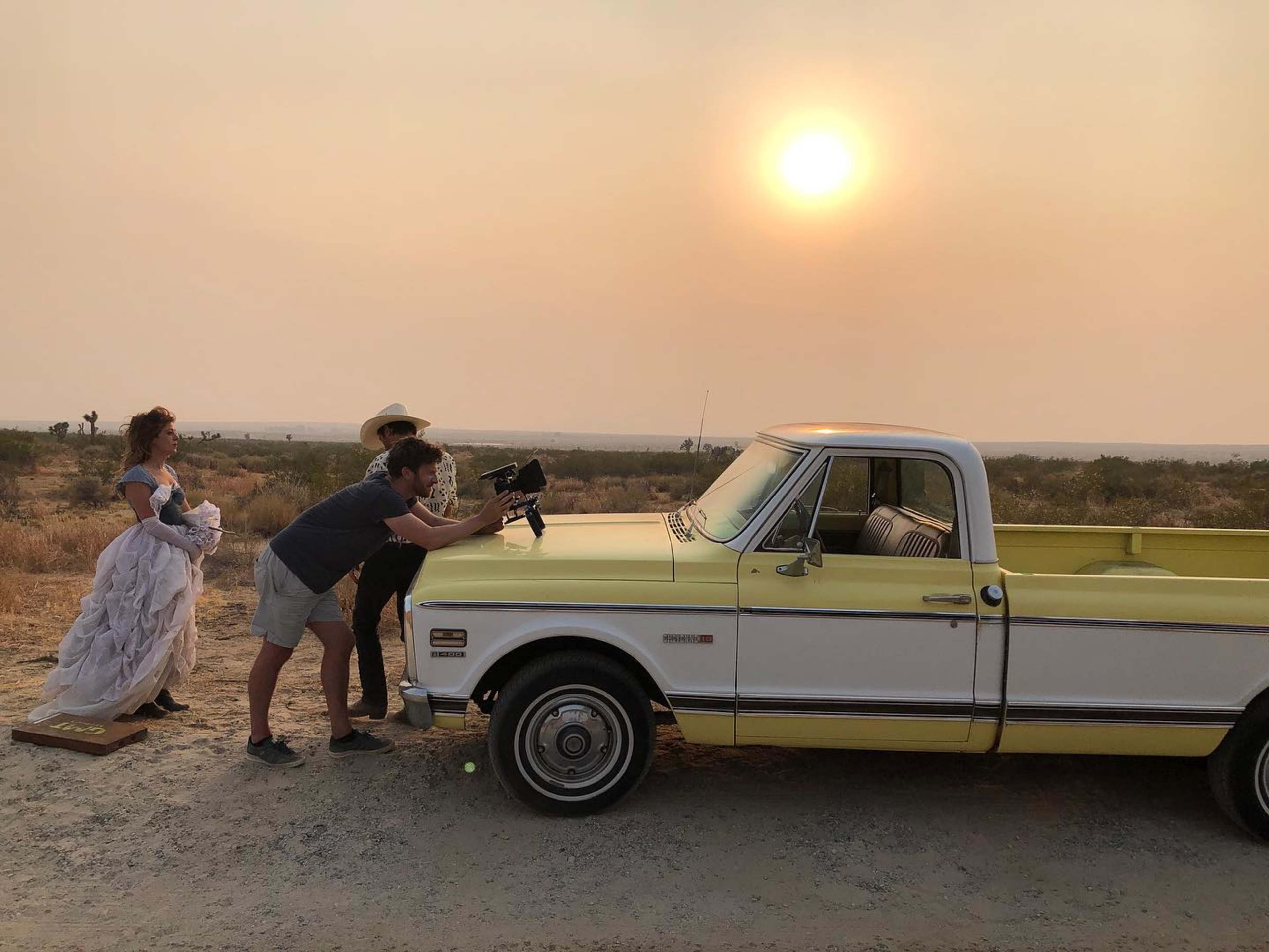 Three people set up a camera in front of a vintage yellow and white pickup truck on a dusty road under a hazy sky with a setting sun.