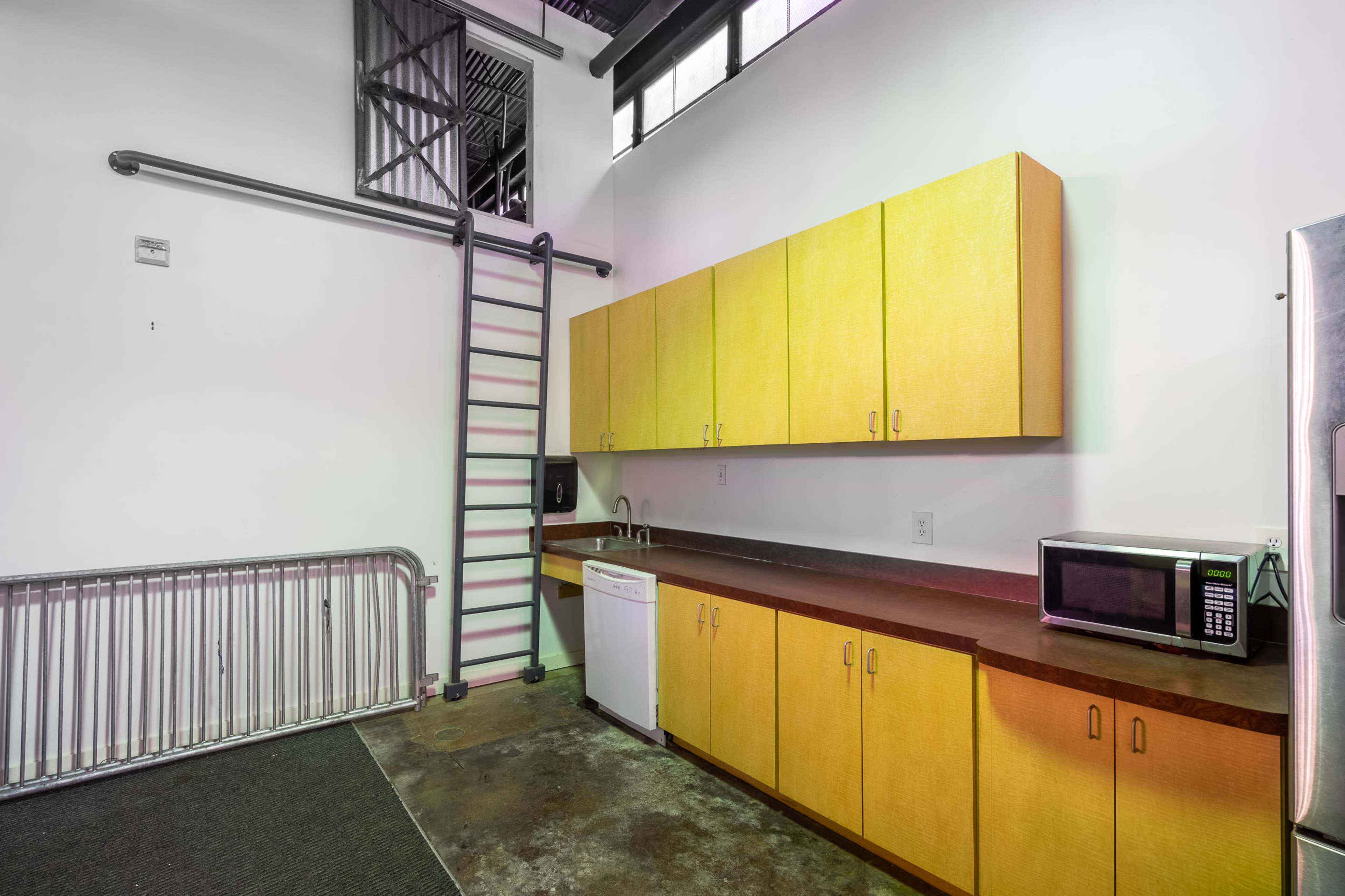 The image shows a minimalist kitchen with yellow cabinetry, a microwave, a dishwasher, and a metal ladder leading to a loft area.