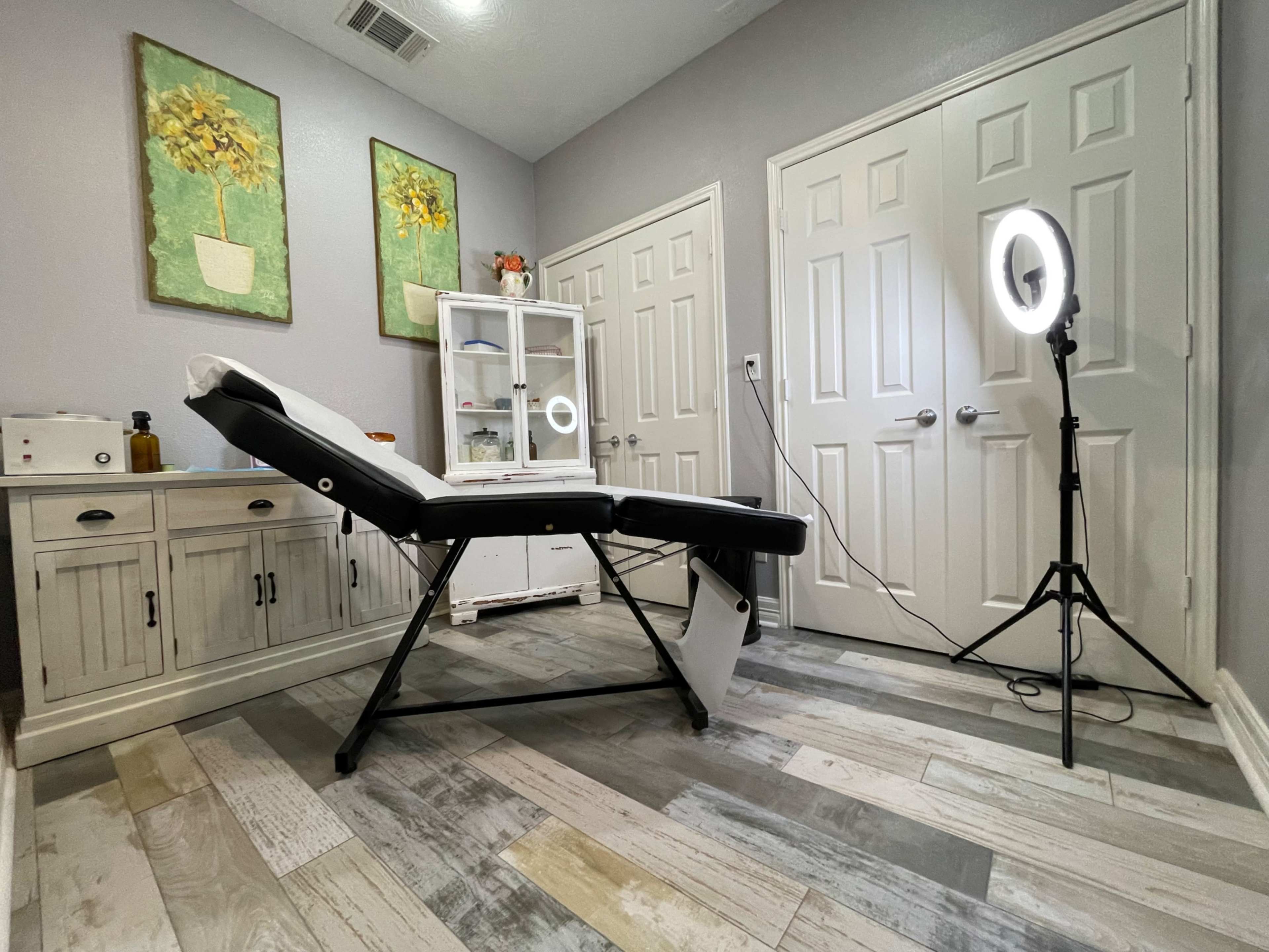 A treatment room with a massage table, a ring light on a tripod, and two closed doors, complemented by two wall-mounted artworks.
