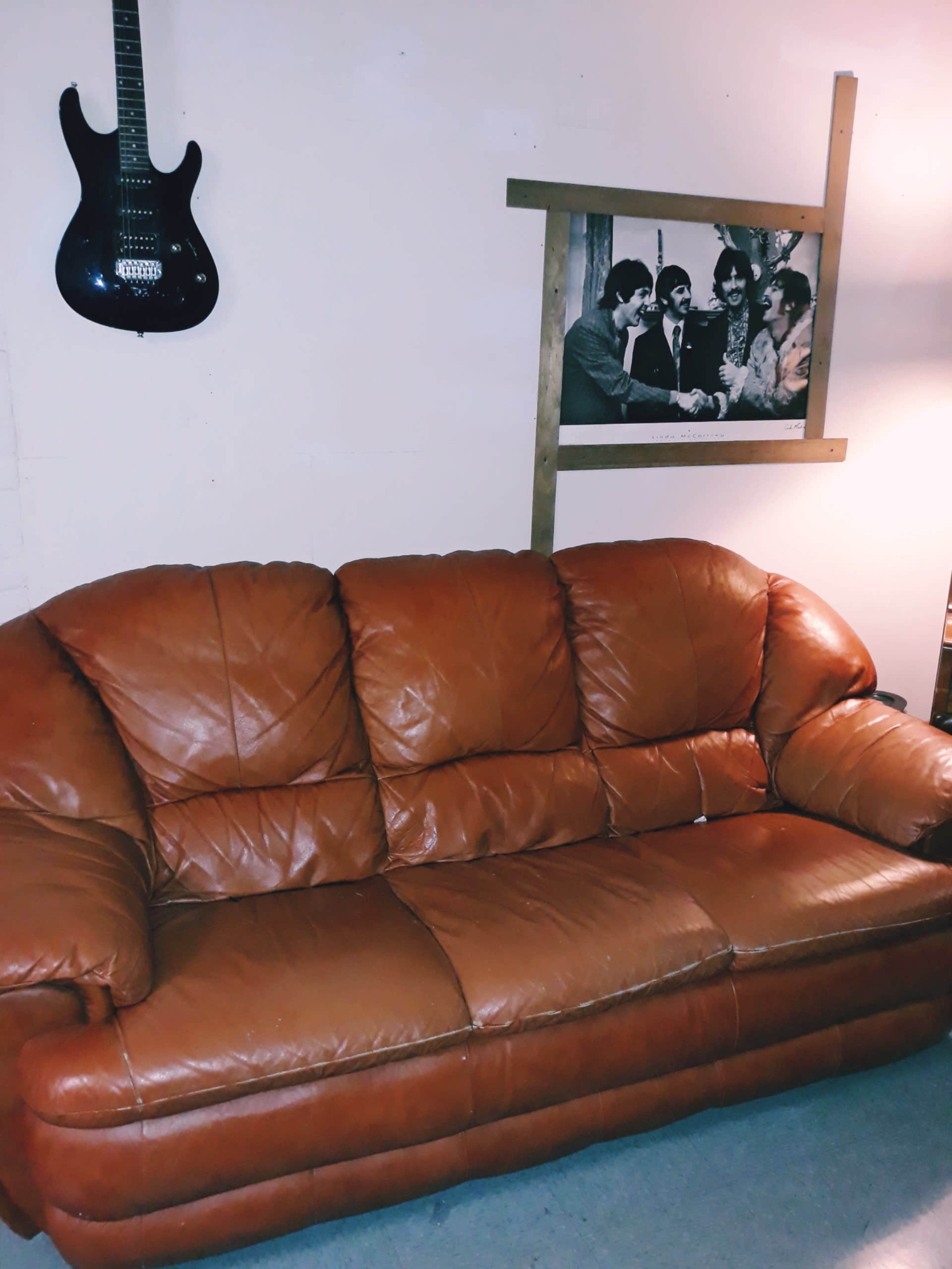 A brown leather couch positioned against a light-colored wall with a framed black-and-white photo of four people on display above it, alongside a black guitar hanging on the wall.