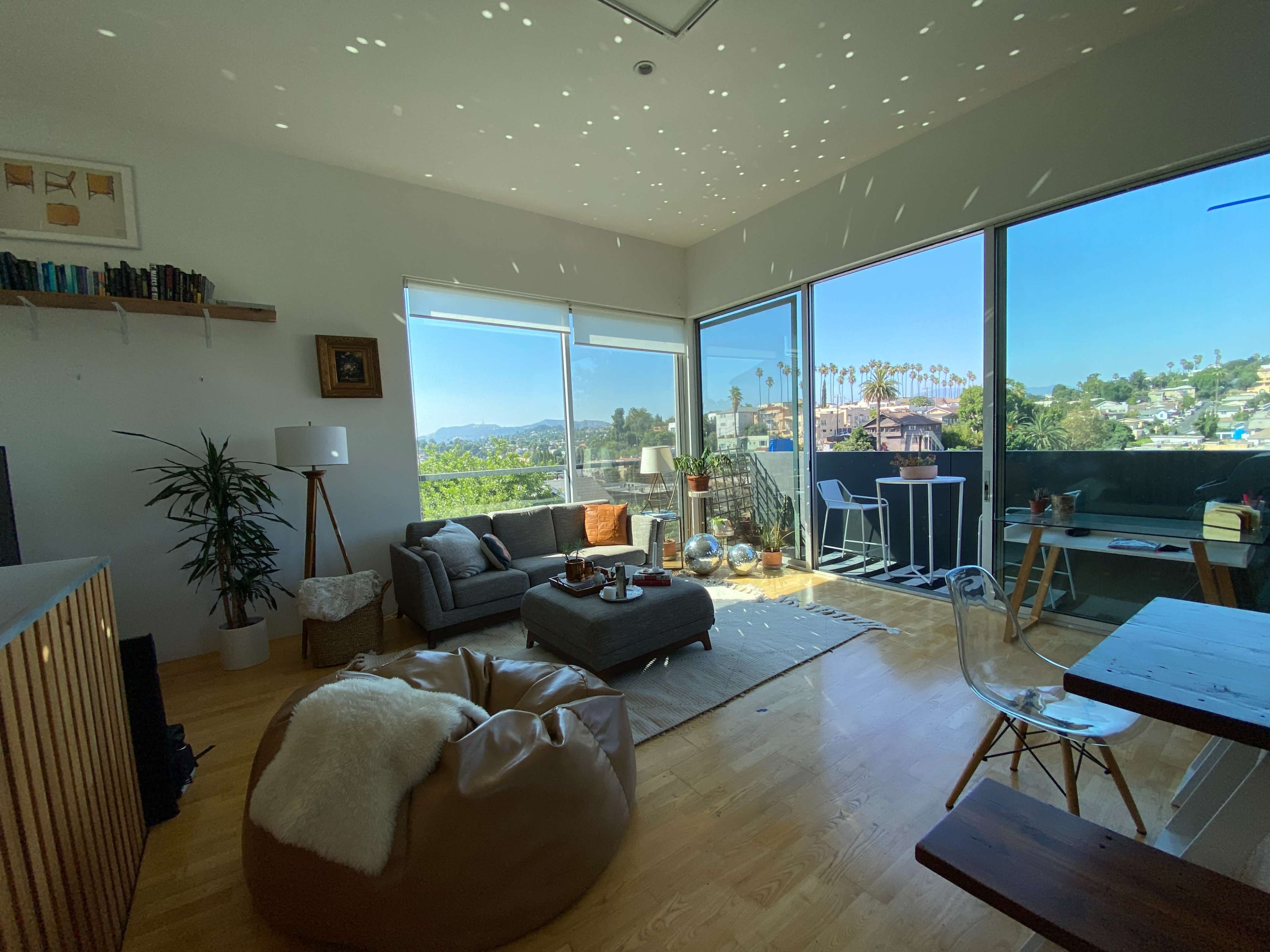 A modern living room features a gray sofa, a large window with a view of palm trees, and light spots dancing on the ceiling.