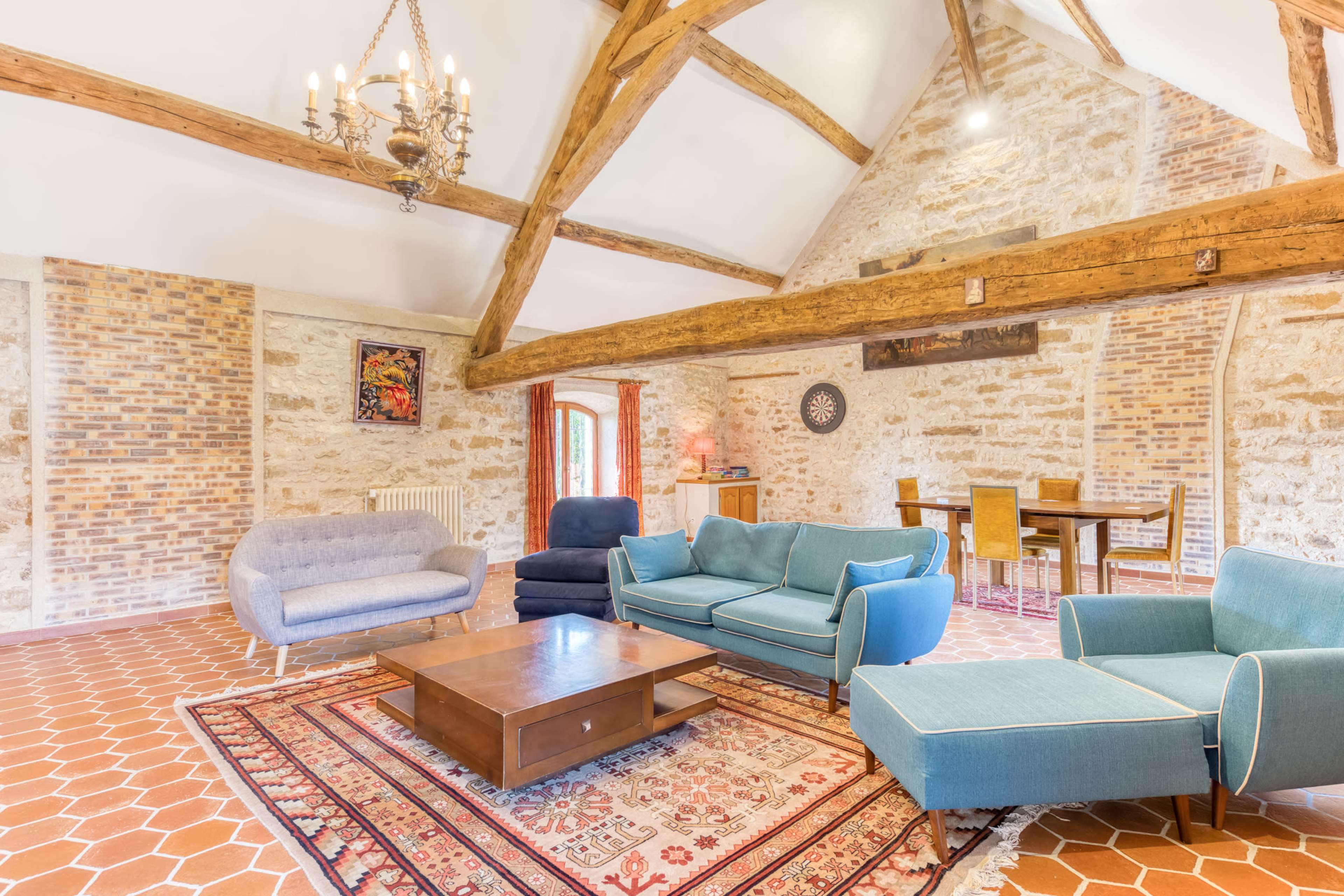 The image shows a spacious living area with a high ceiling, exposed wooden beams, and a mix of comfortable seating arrangements, including a couple of sofas and a lounge chair, alongside a wooden dining table in the background.