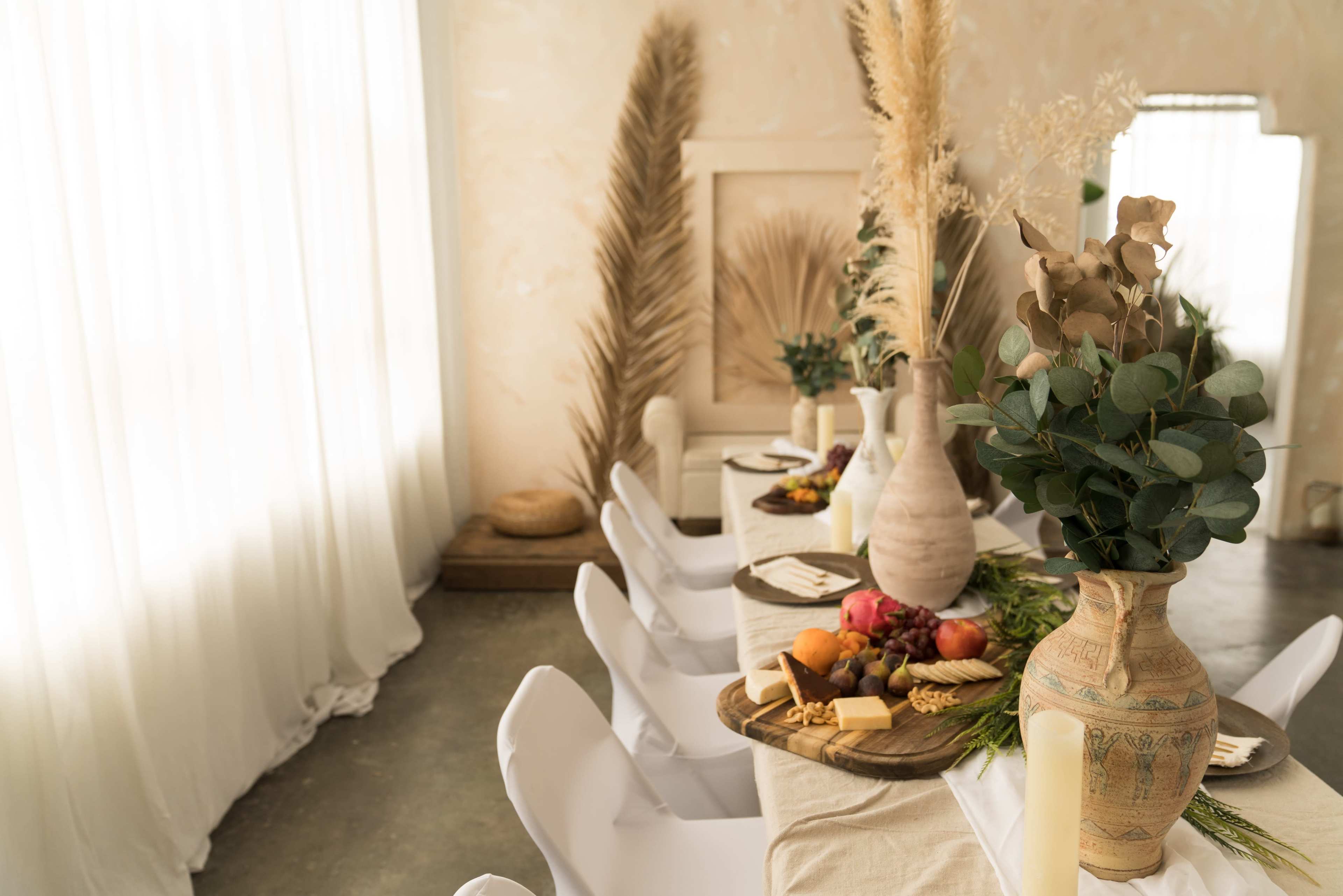 A dining table is set with a variety of fruits, cheese, and decorative elements, surrounded by white chairs in a softly lit indoor space.