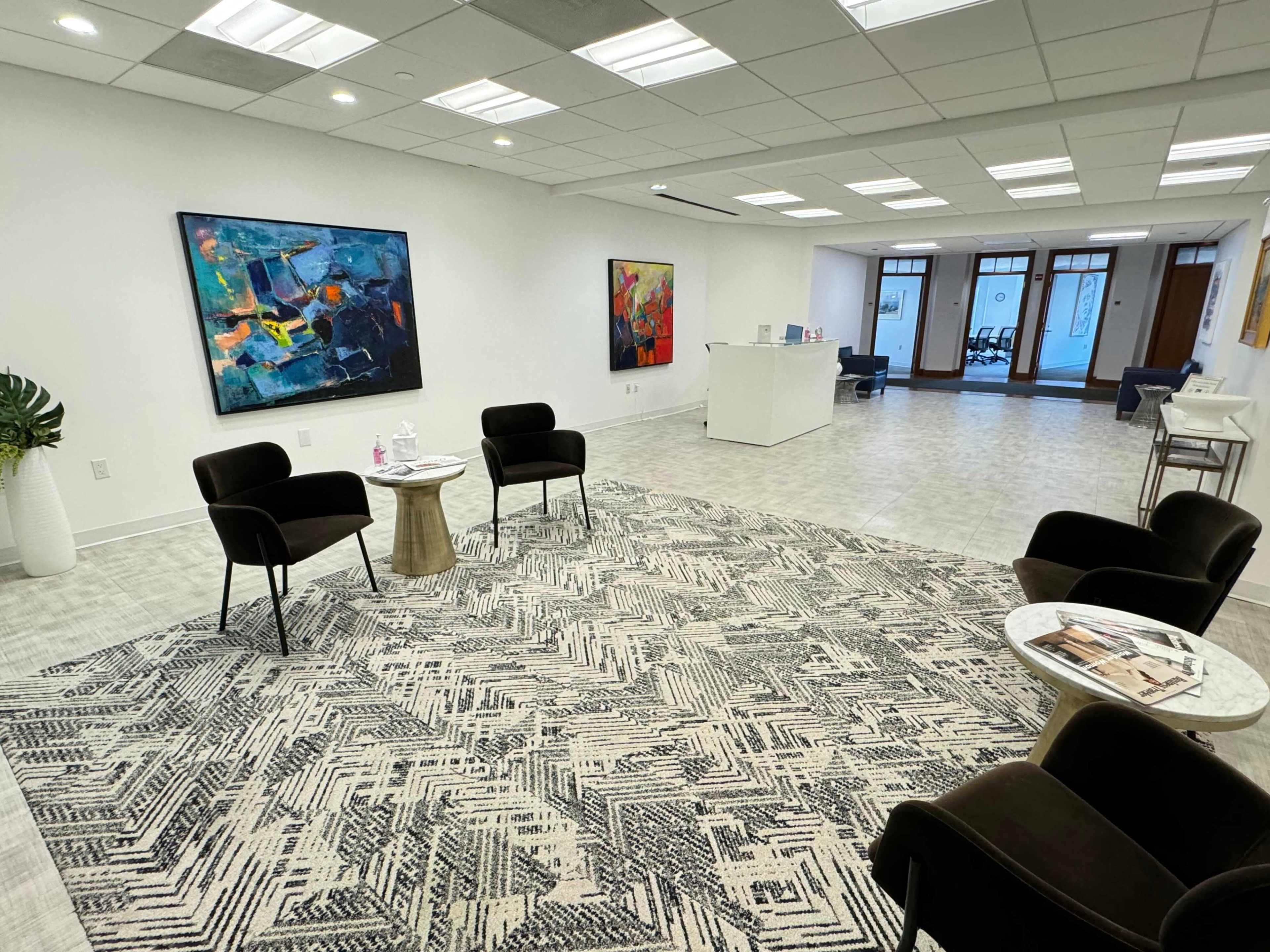 The image shows a modern office space with two abstract paintings on the walls, a patterned rug on the floor, and several black chairs arranged around a small table.