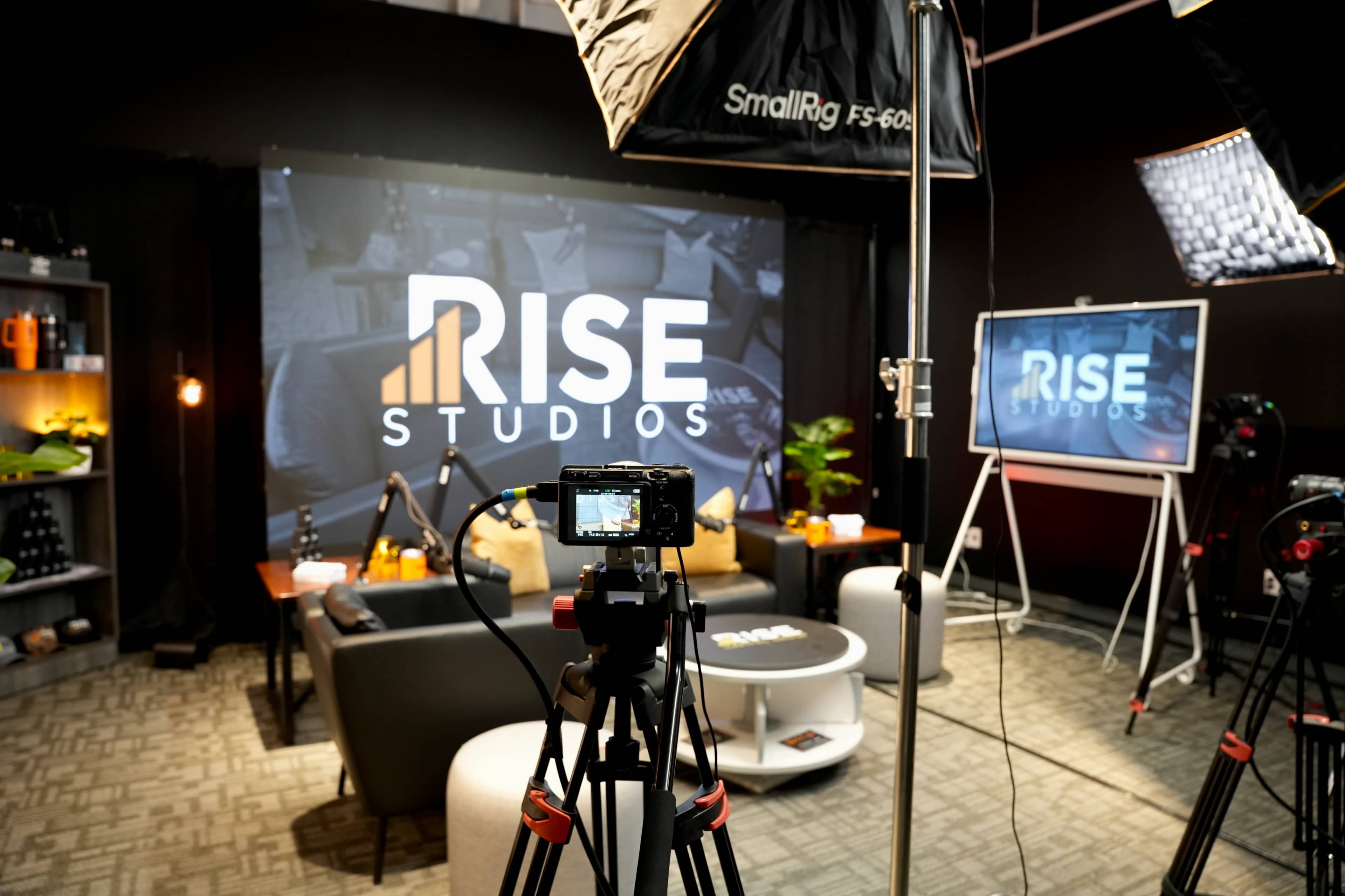 The image shows a television studio setup featuring a logo for "RISE STUDIOS," with cameras, lighting equipment, and a modern seating arrangement.