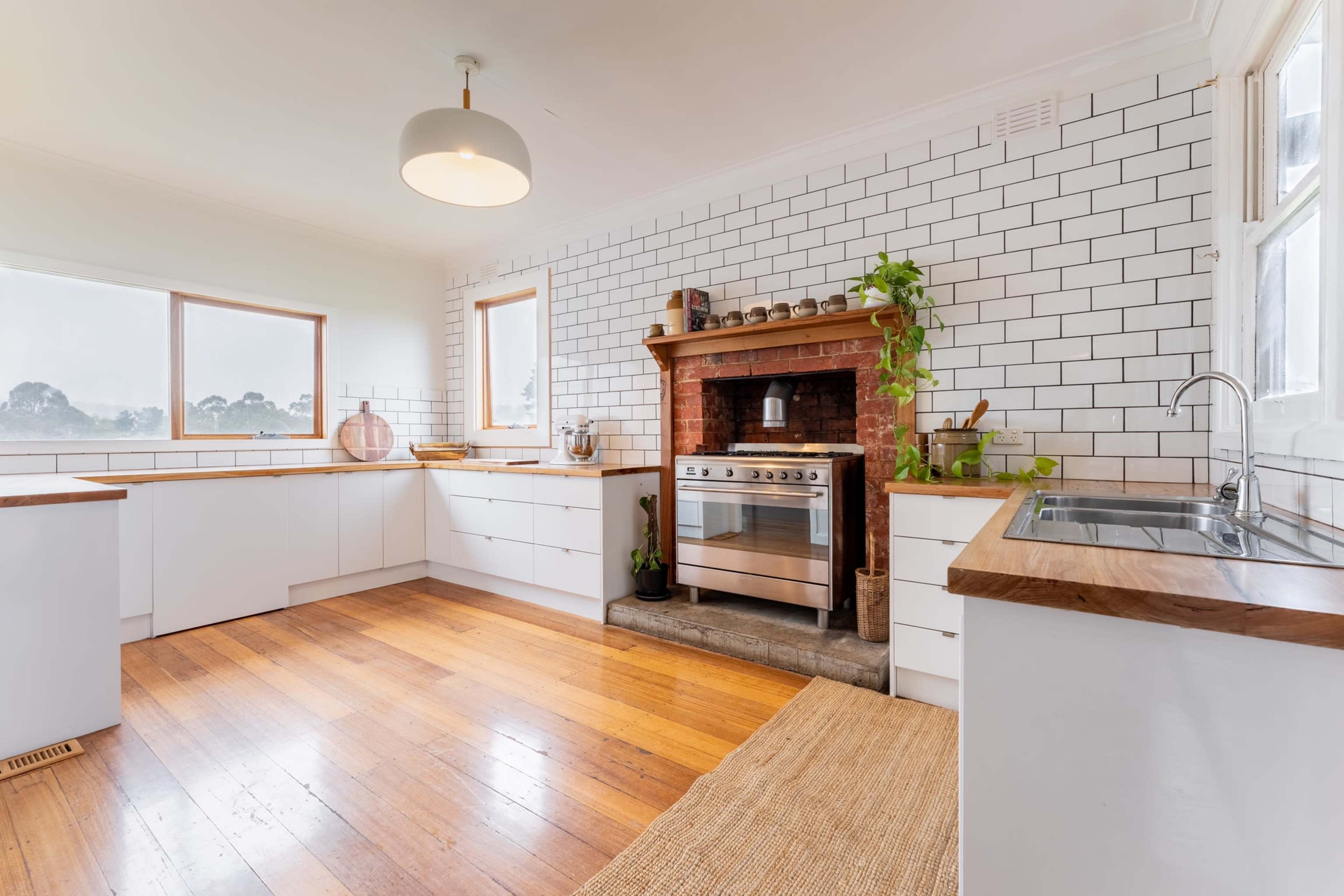 A modern kitchen features white cabinetry, a brick fireplace with an oven, and wooden floors, illuminated by natural light through large windows.