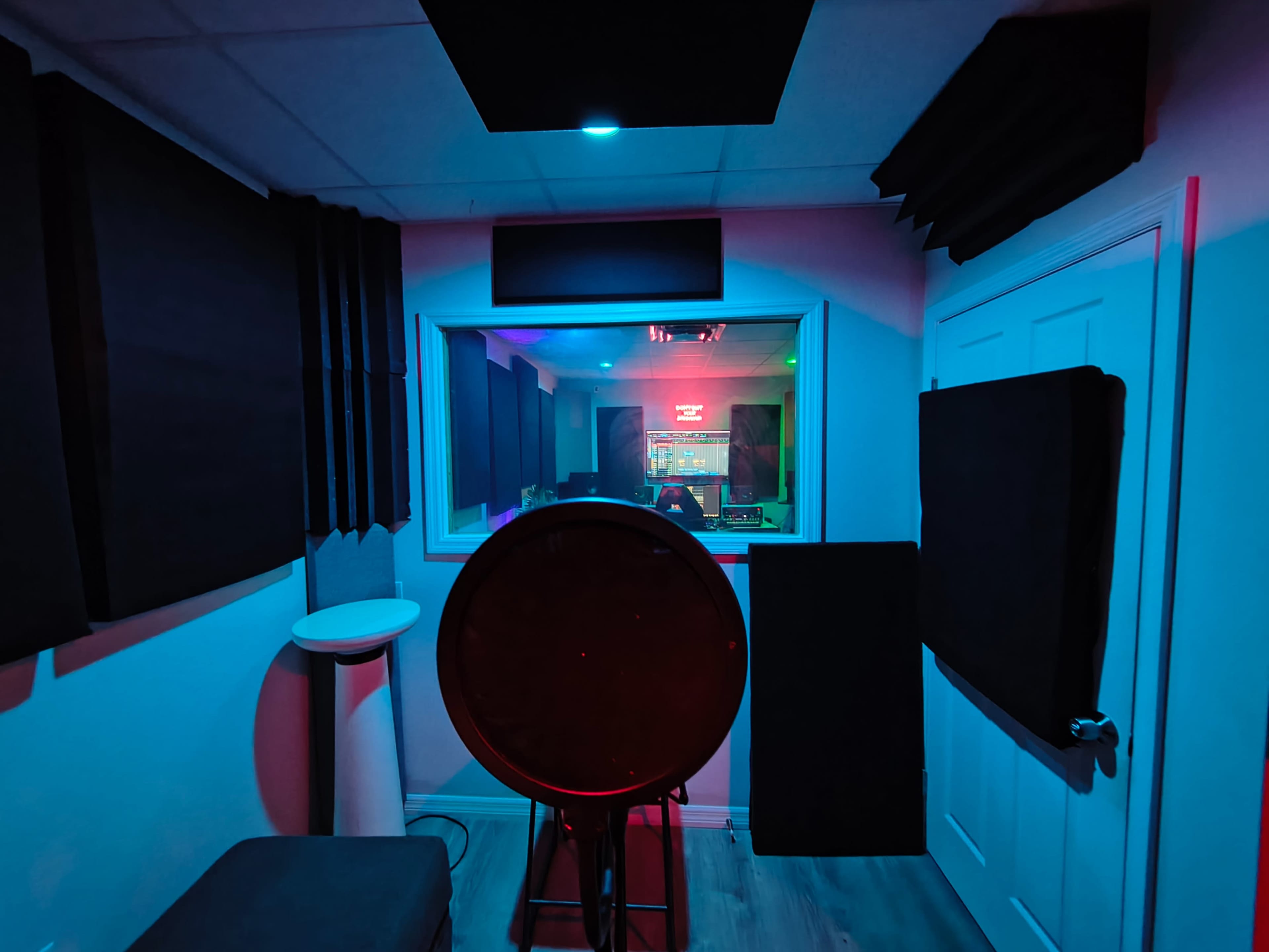 The image shows a soundproof recording studio with acoustic panels on the walls, a circular microphone setup in the foreground, and a control room visible through a glass window in the background.