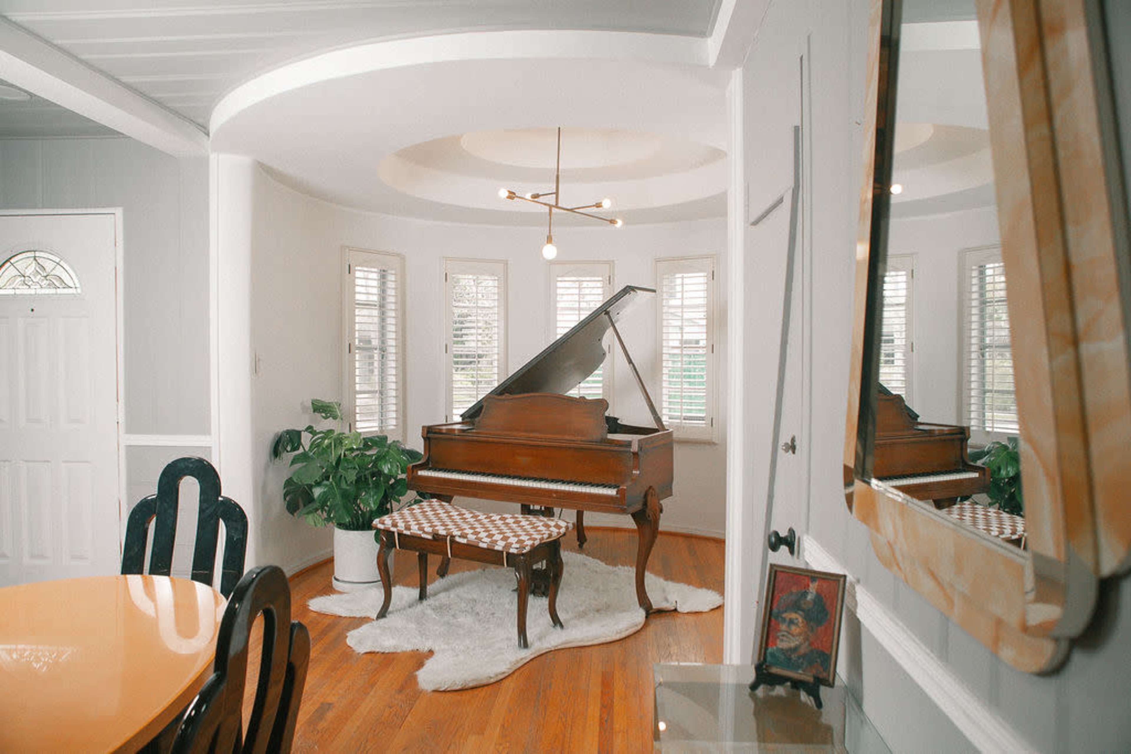 A grand piano sits in a brightly lit corner of a room, accompanied by a bench and surrounded by windows with plantation shutters.