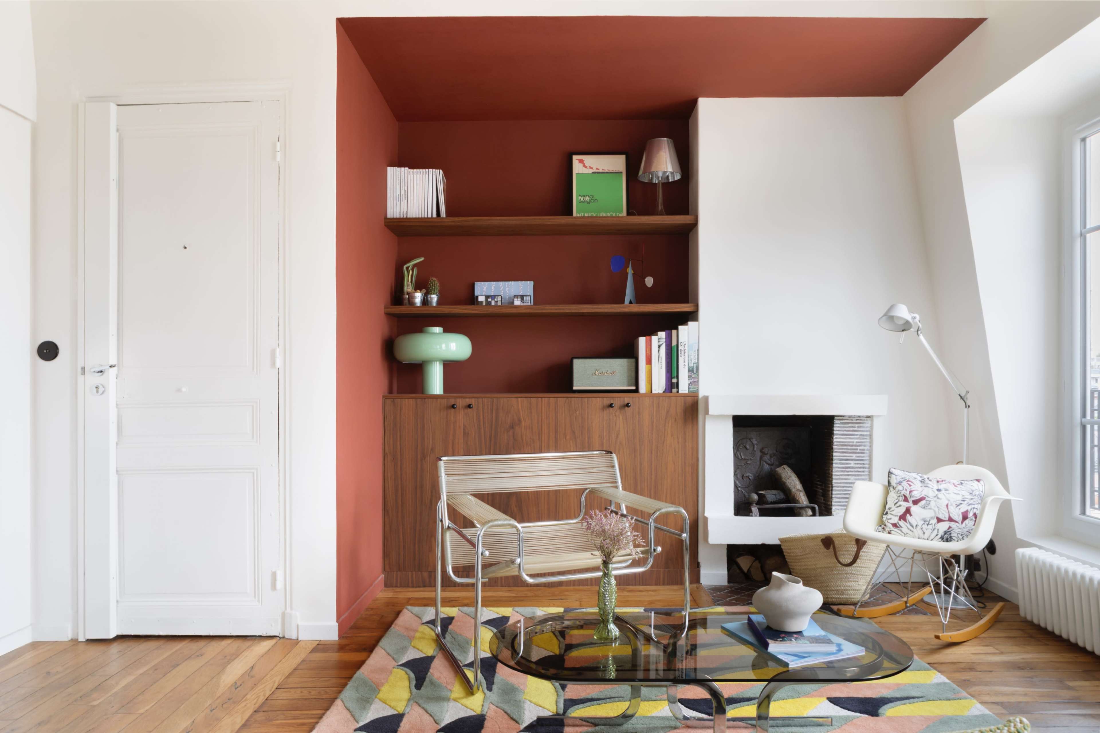The image depicts a modern living room featuring a minimalist glass coffee table, a stylish armchair, and a wooden shelf with books and decorative items against a red accent wall.