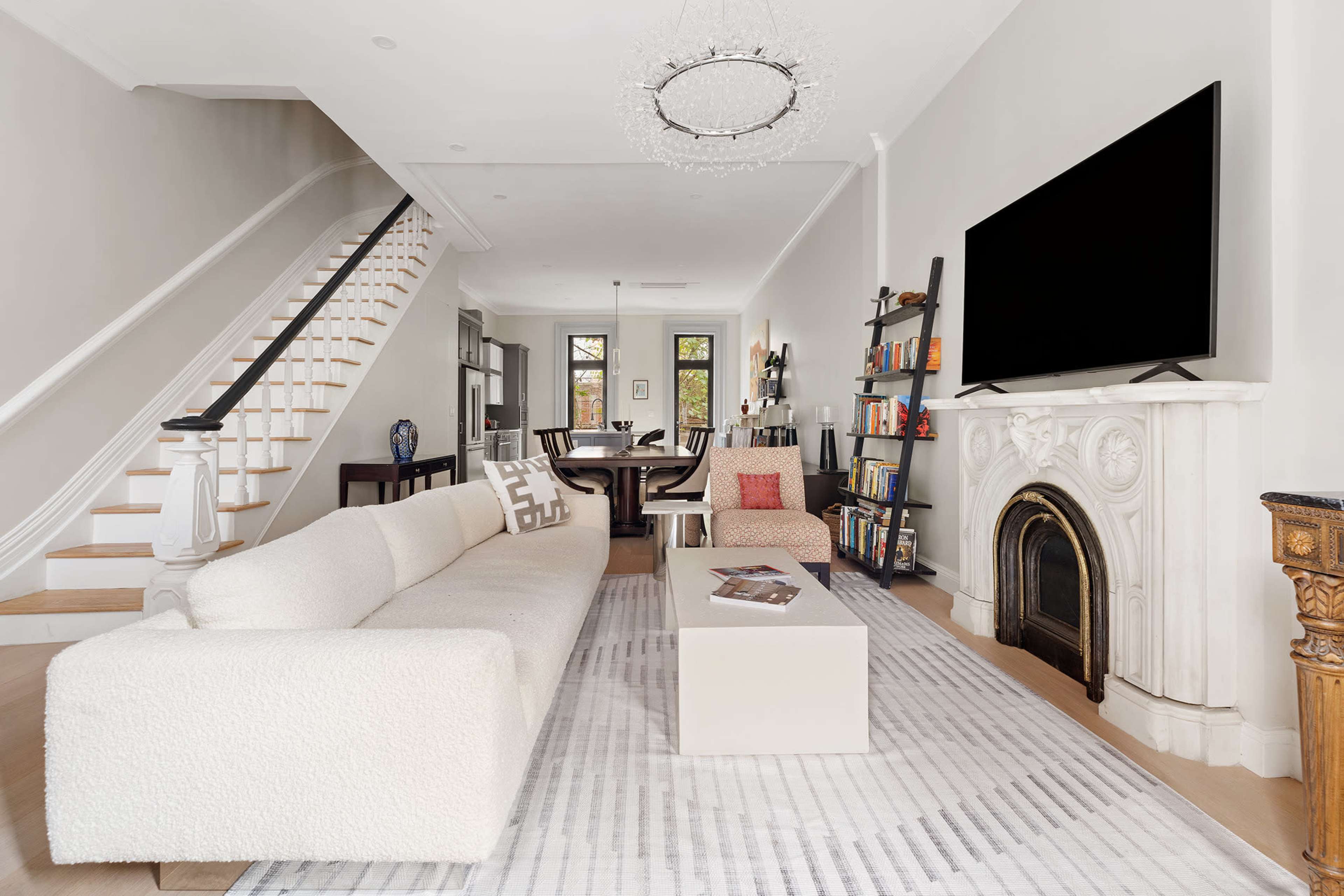A modern living room features a large, light-colored sofa, a coffee table, a flat-screen TV mounted on the wall, a decorative fireplace, and a staircase leading to the upper level.