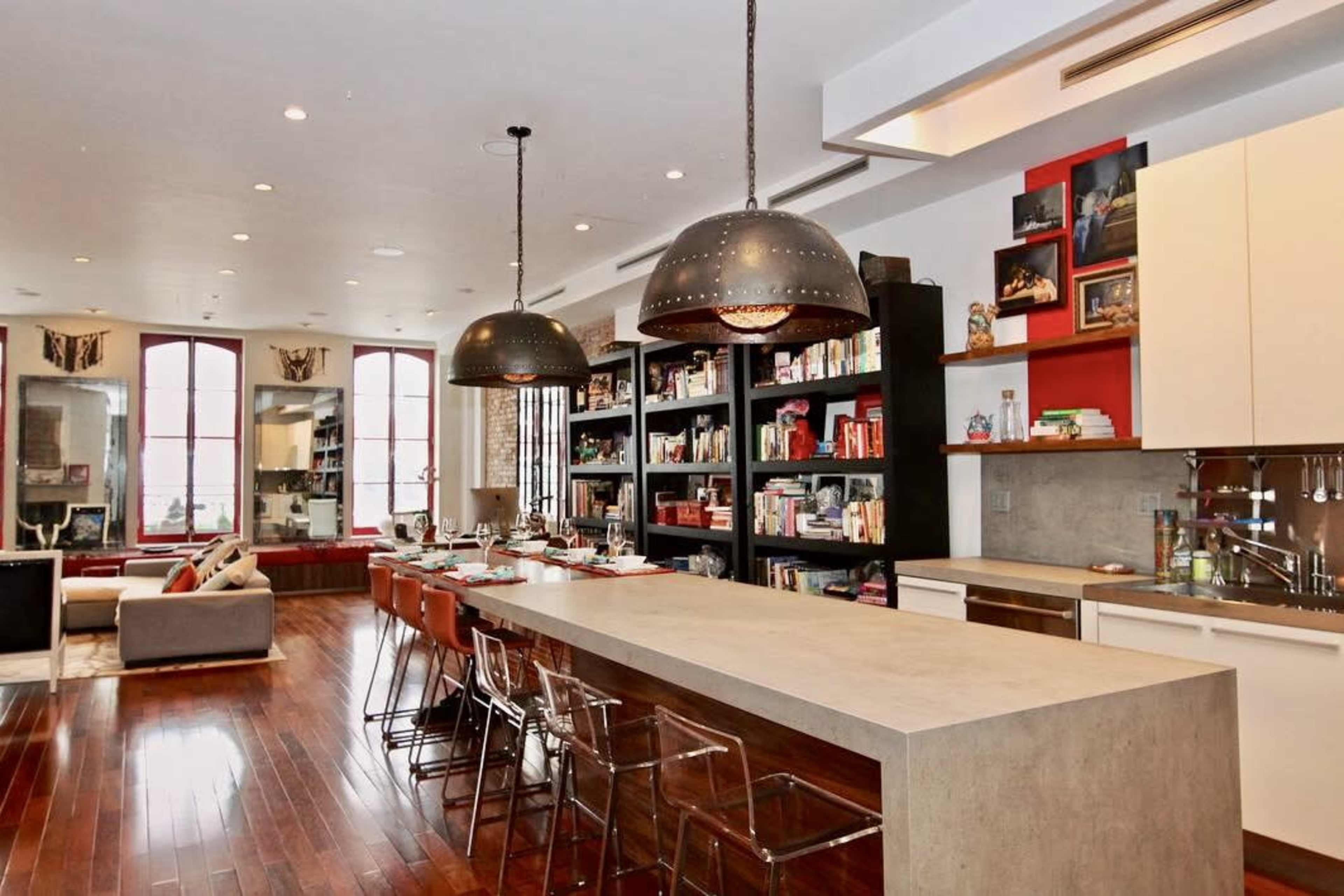 The image shows a modern kitchen with a long concrete island, bar stools, and dark wooden flooring, accompanied by shelves filled with books and decorative items.