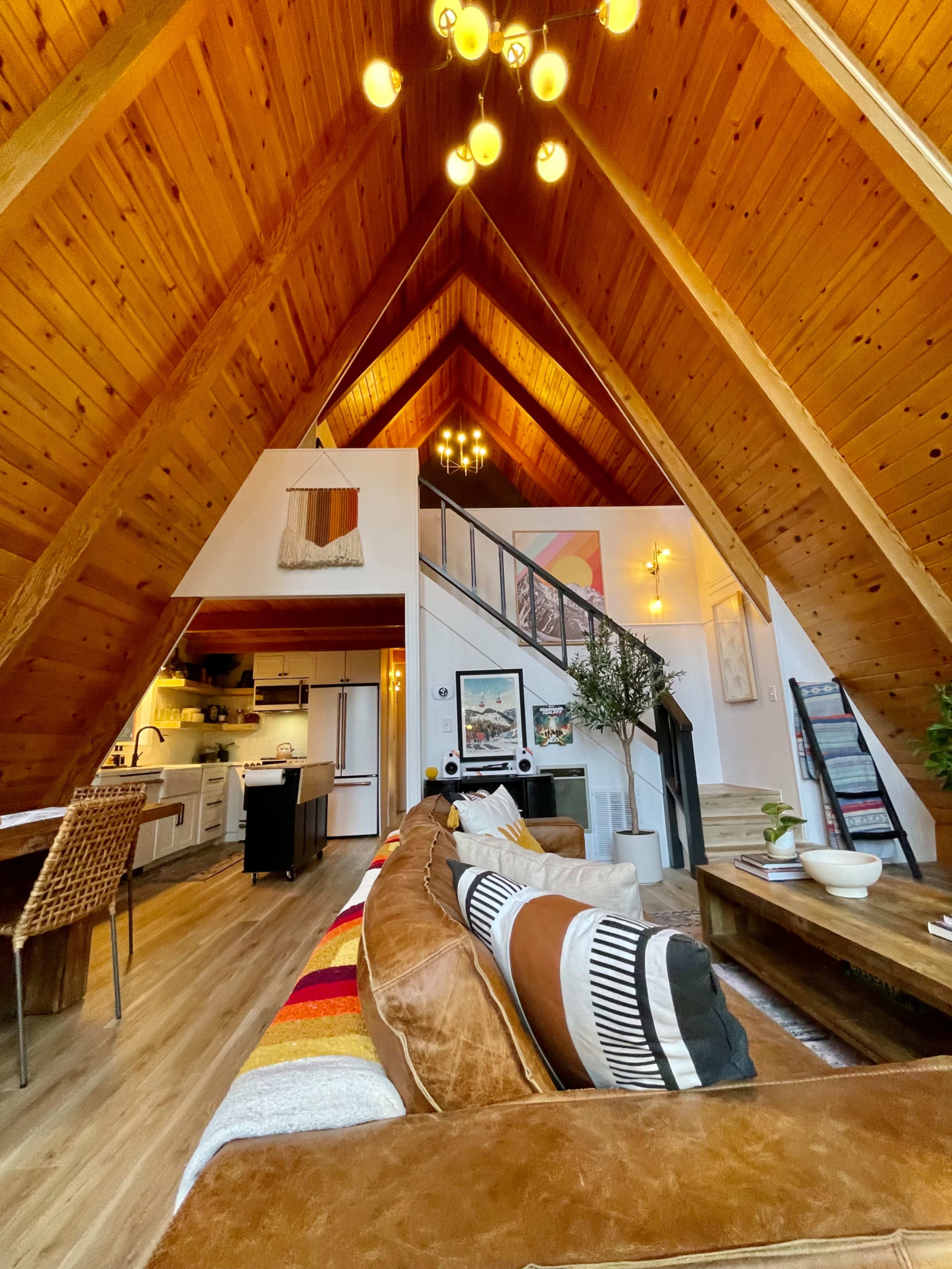 The interior of a modern A-frame cabin features high wooden ceilings, a cozy living area with a sectional sofa, and a dining space visible in the background.