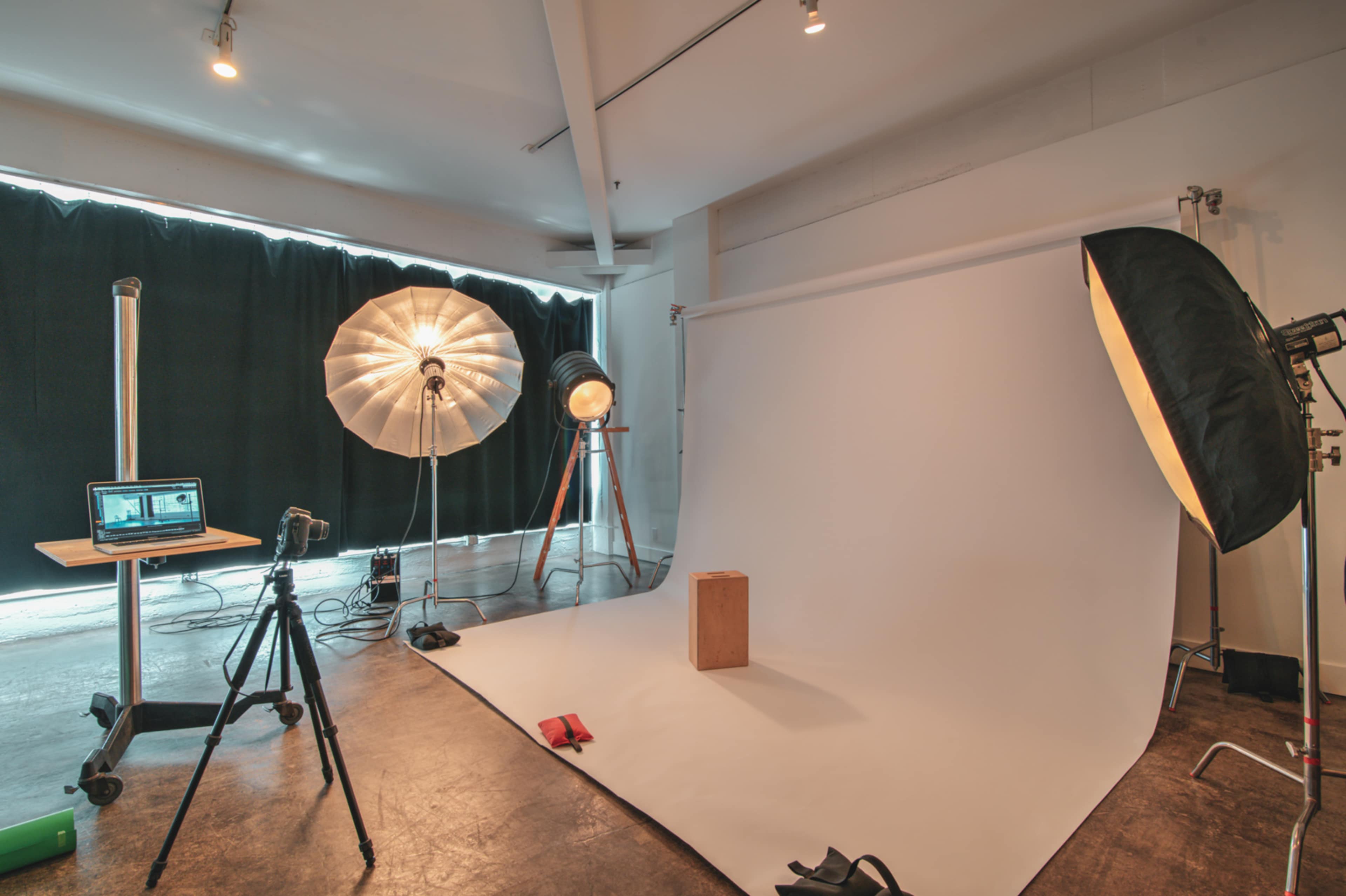 A photography studio setup with a seamless backdrop, a light stand, softboxes, and a tripod positioned near a wooden block.
