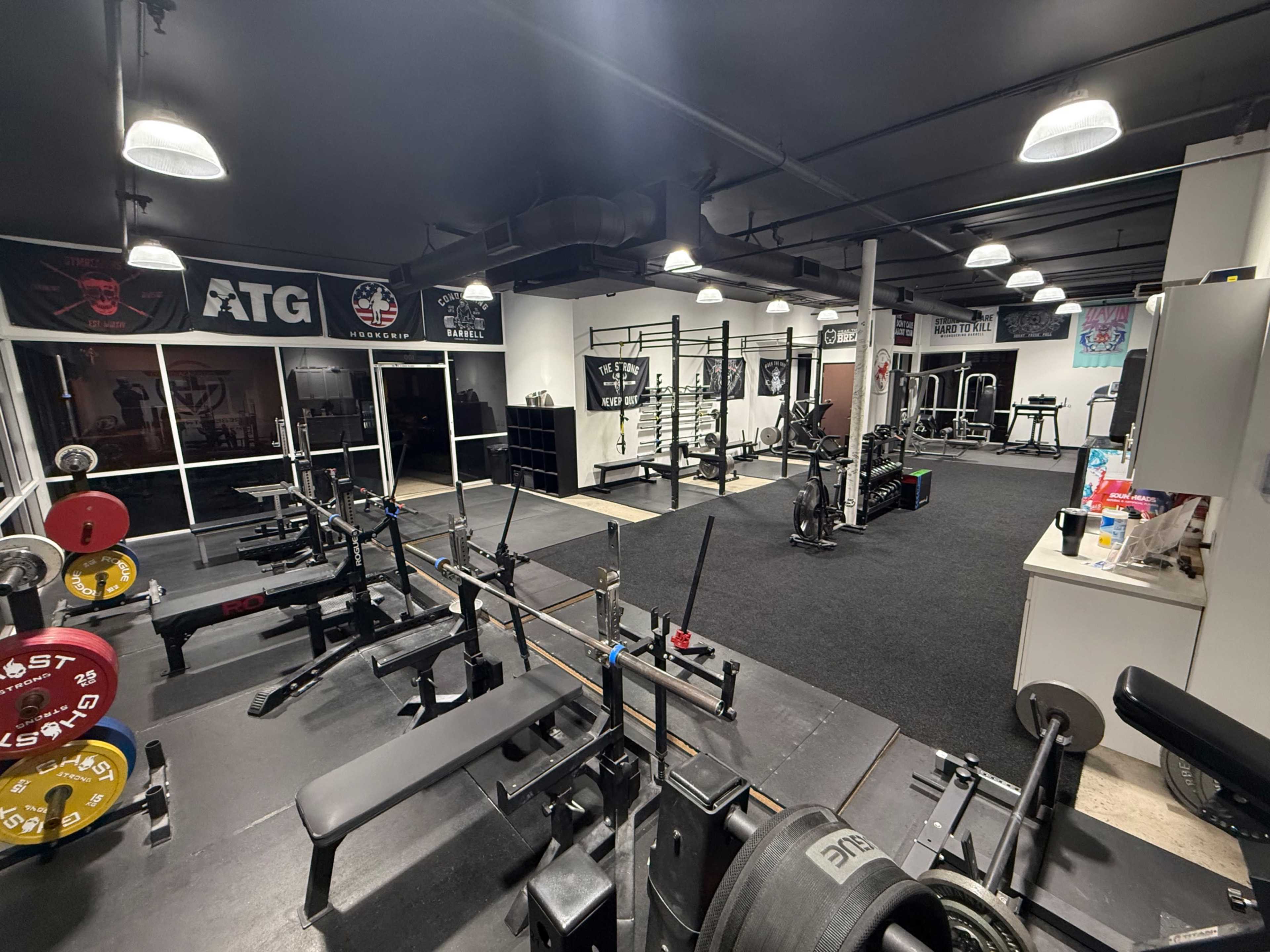 The image shows an indoor gym space equipped with various weightlifting machines, benches, and free weights, along with wall posters and a large window.