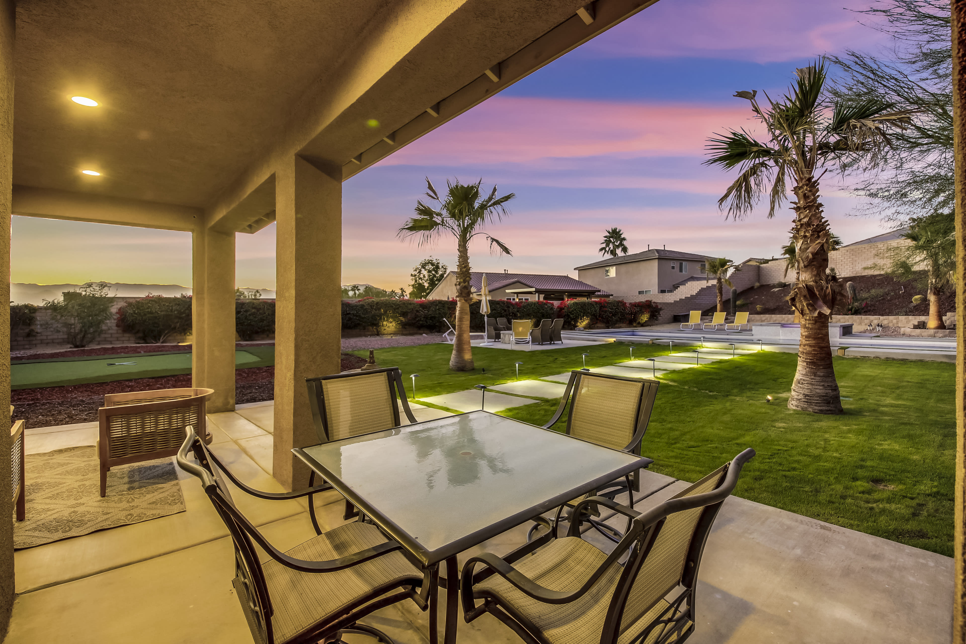 A glass table with metal chairs is set on a patio overlooking a landscaped yard featuring palm trees and lounge chairs against a colorful sunset sky.