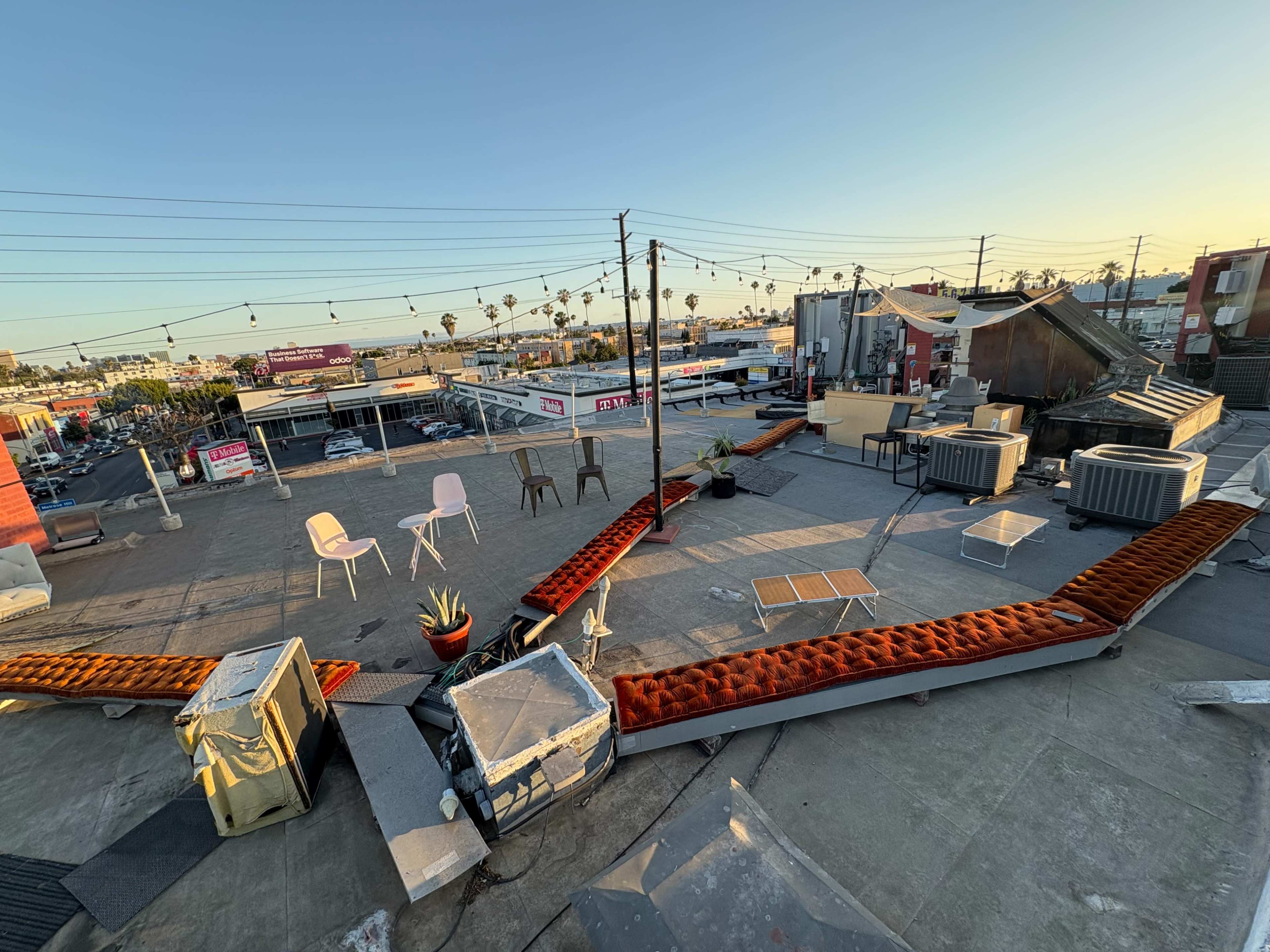 The image shows a rooftop patio area with scattered furniture, including chairs and a table, overlooking a bustling street with businesses and palm trees in the background.