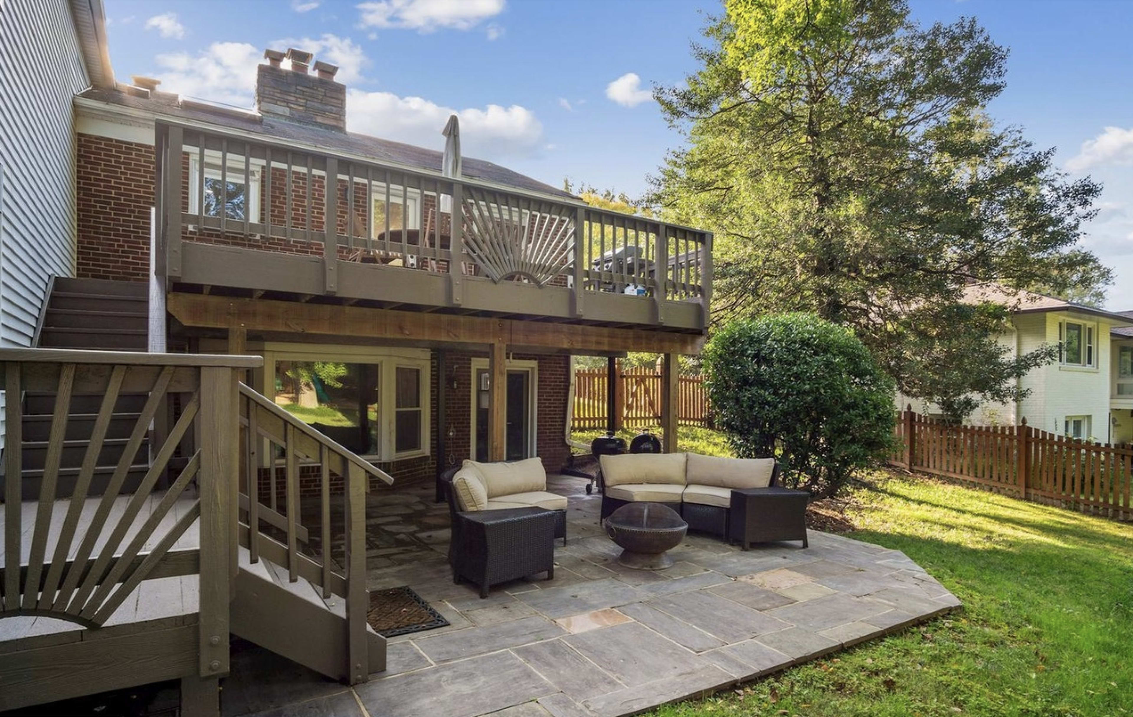 The image shows a backyard with a stone patio, outdoor seating, and a wooden deck above, surrounded by trees and a fence.
