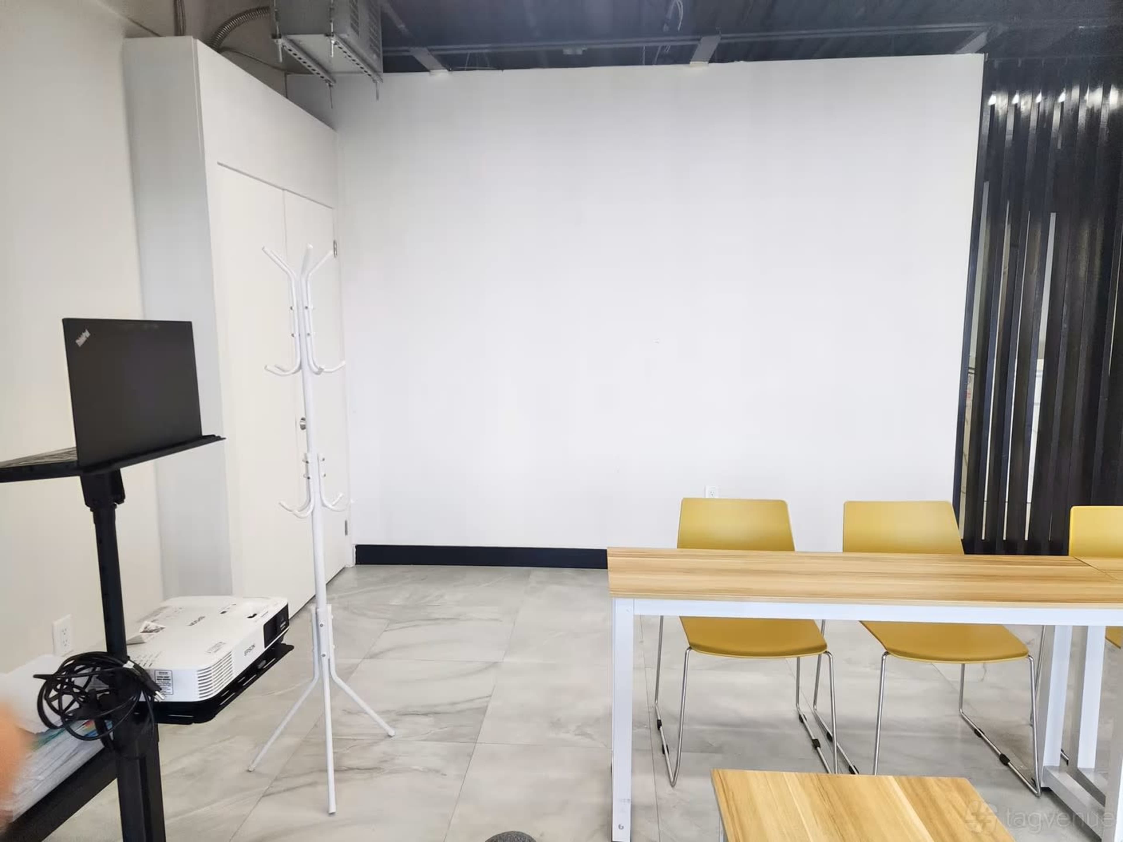 The image shows a minimalistic room with a projector, a laptop stand, a coat rack, and a table with four yellow chairs, set against a plain white wall.