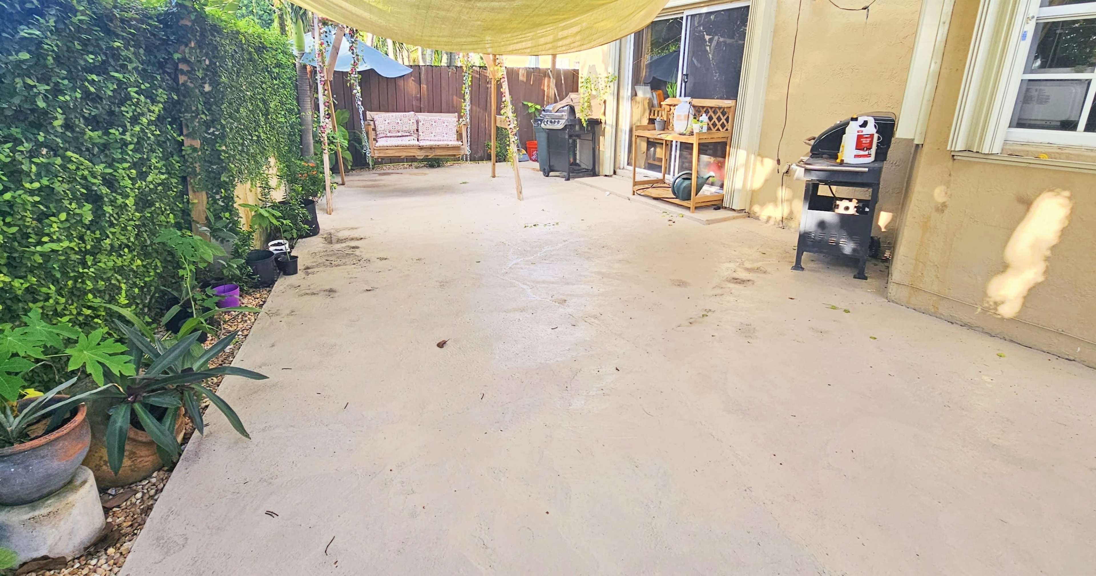A spacious outdoor area with a concrete surface, a shaded seating area with a couch in the background, and a barbecue grill to the right.